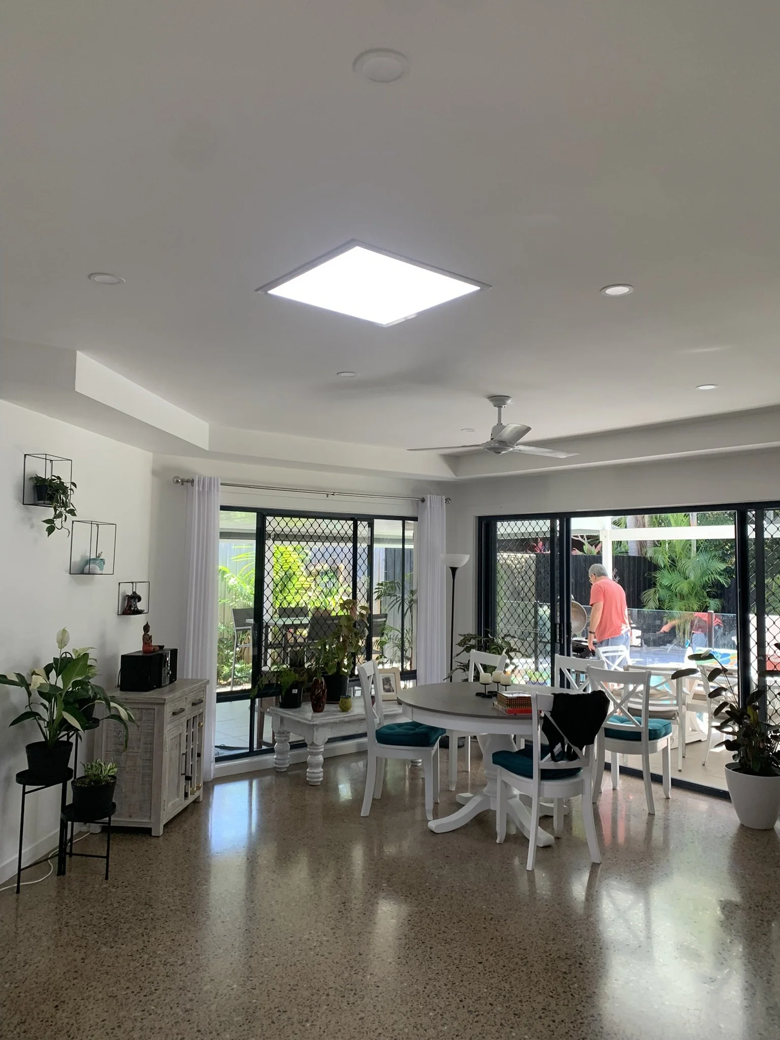 Bright living room lit up by an illume skylight with plants and round dining table near large sliding glass doors, leading to a patio with a man outside.