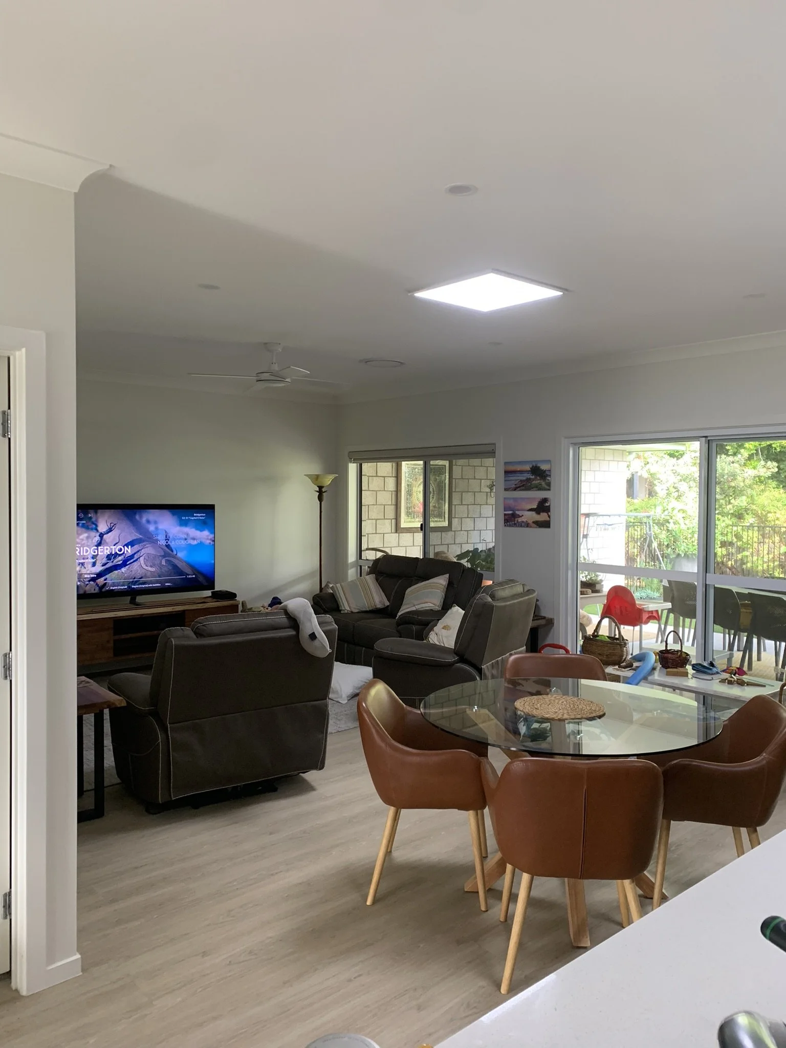 A modern living room with a glass dining table and brown chairs, a TV, and cozy seating area. The room has light wooden flooring and is well-lit by an illume skylight, with sliding doors leading to an outdoor area.