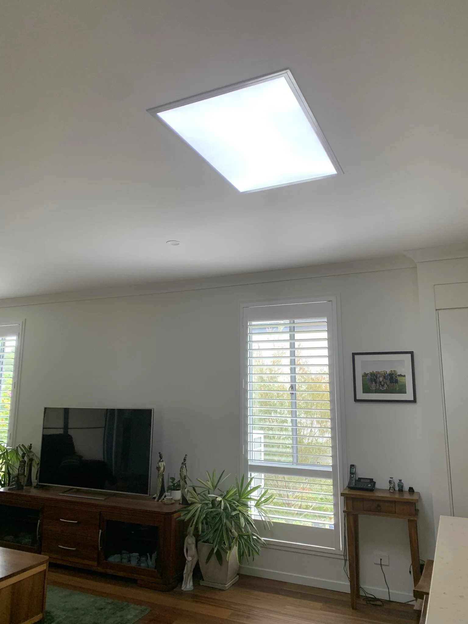 Interior of a room with a ceiling light, television on a wooden cabinet, windows with blinds, potted plants, and framed wall artwork. The illume skylight bring a great feature into the room.
