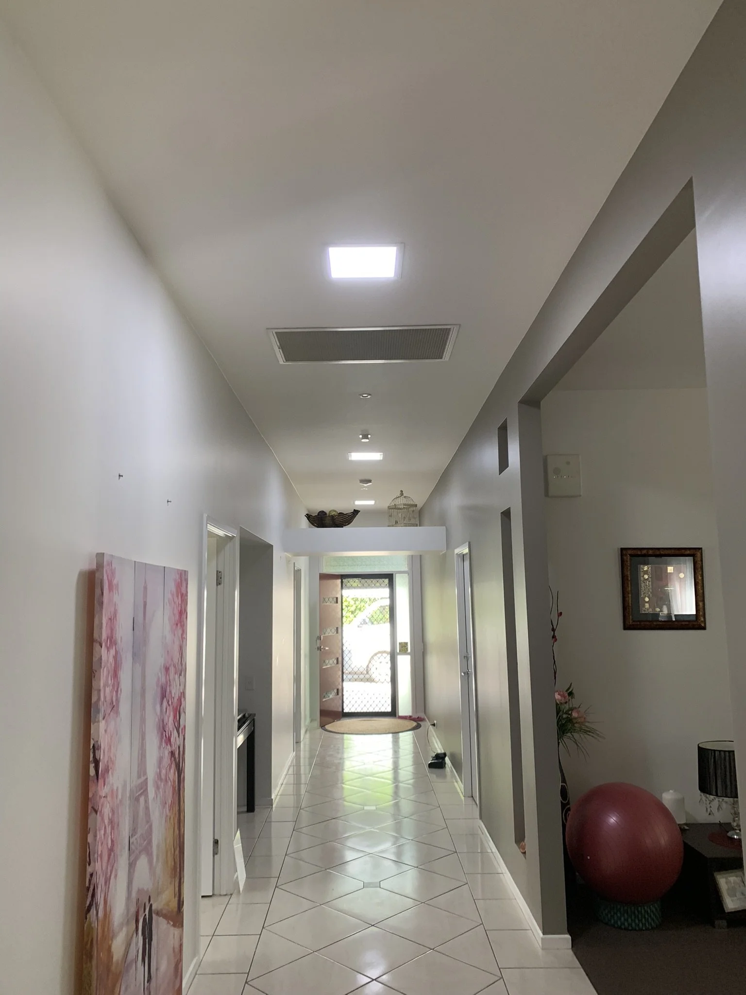 Interior hallway with white walls, illume skylights, tiled floor, a yoga ball, framed artwork, and a glass door at the end.