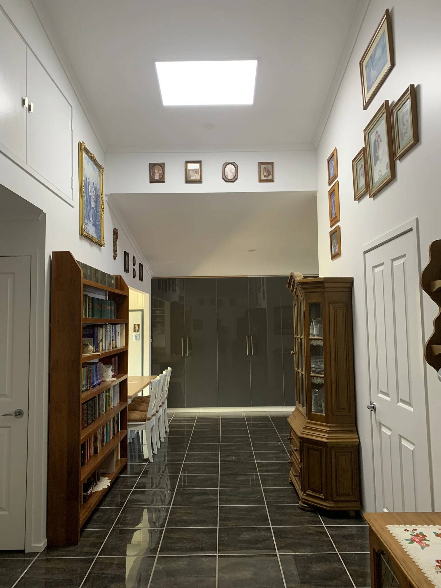 Interior hallway with black tiled floor, wooden bookshelf filled with books on the left, wooden cabinet on the right, table with chairs in the background, and family photos or artwork on the walls. A large solar skylight illuminate the space.