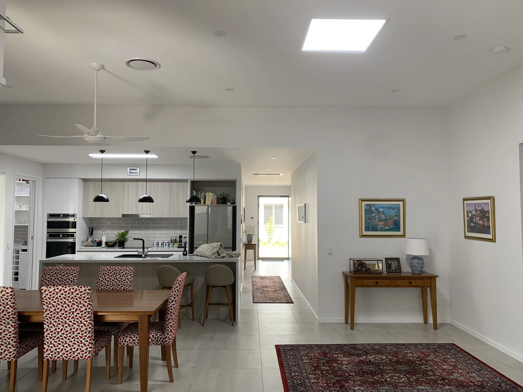 Modern open-plan kitchen and dining area with white walls, tiled floor, and a ceiling fan. The kitchen features a large island with bar stools, illume skylights, and stainless steel appliances. A wooden dining table with patterned chairs is nearby.