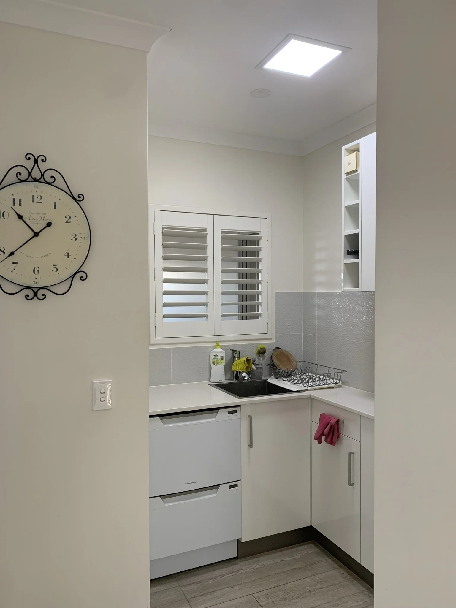 Small modern kitchen with a white counter, double dishwasher, black sink, white cabinets, wall clock, and window with shutters. A pink towel hangs from a cabinet handle. illume skylight makes the space radiant. 