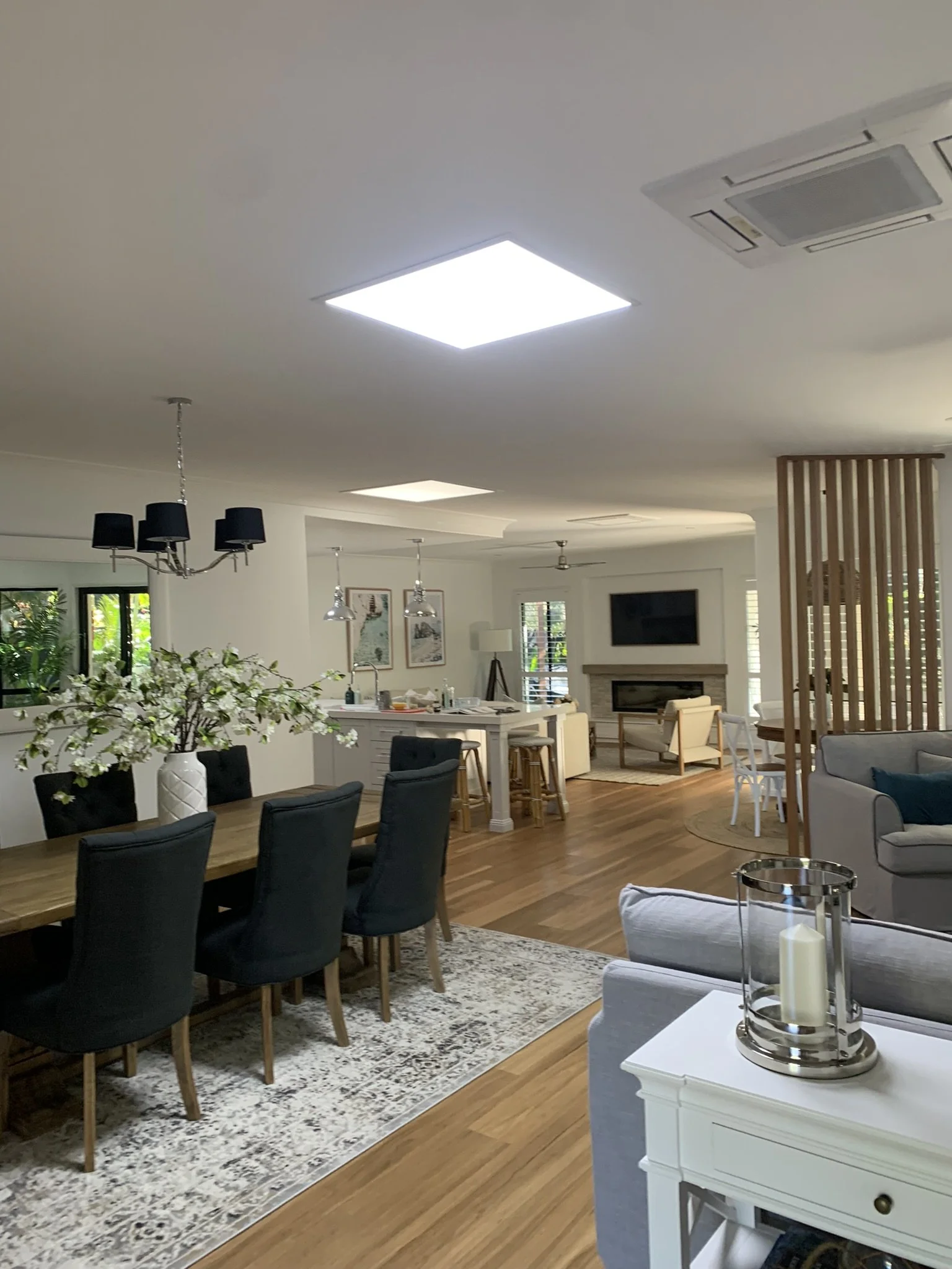 Open-concept living and dining area with illume skylight, wooden floors, a large dining table, black dining chairs, a gray sofa, and a modern kitchen. Interior decorated with a centerpiece of white flowers and a candle holder on a side table.
