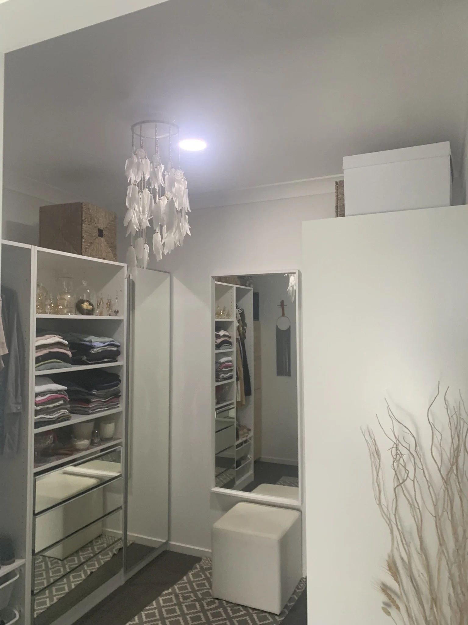 Walk-in closet with organized shelves of folded clothes, a full-length mirror, a white ottoman, white hanging decor from the ceiling, and decorative boxes and vases on top of the shelves. An illume skylight bright up the space.