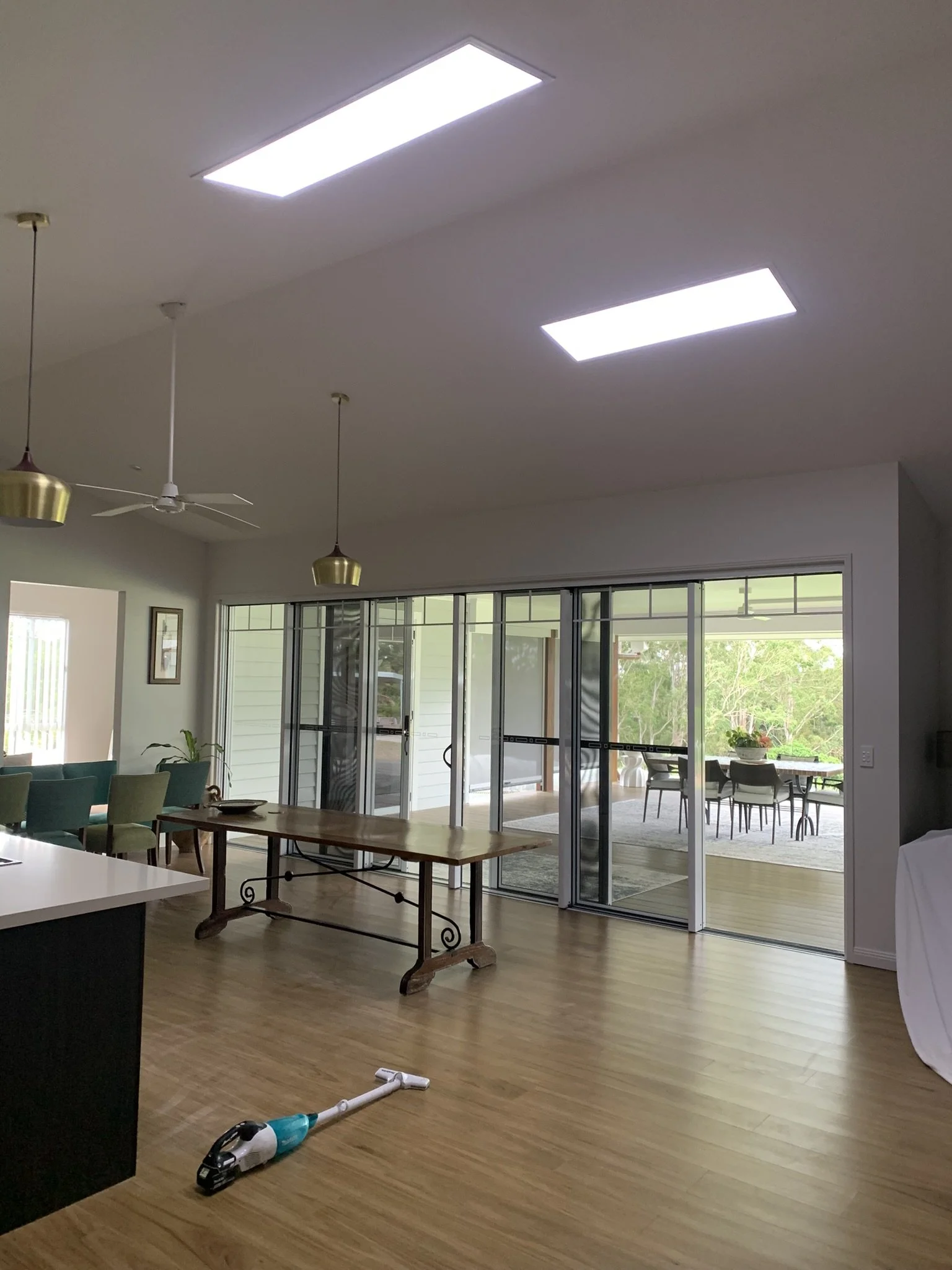 Modern dining room with two illume skylights, wooden flooring, a table, and sliding glass doors opening to a patio. A vacuum cleaner is on the floor, and ceiling lights are visible overhead.