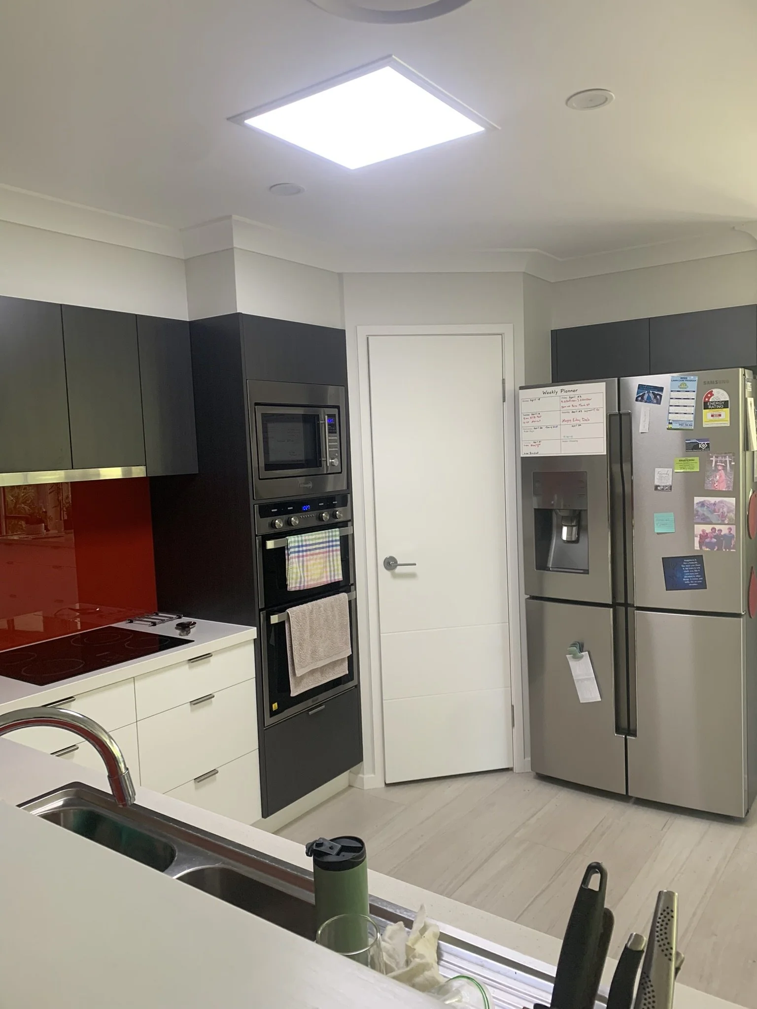 Modern kitchen with stainless steel fridge, built-in oven, microwave, and induction cooktop. The fridge has magnets and a whiteboard planner. The sink area has a faucet with drying utensils. The illume solar skylight is bighting up the space.