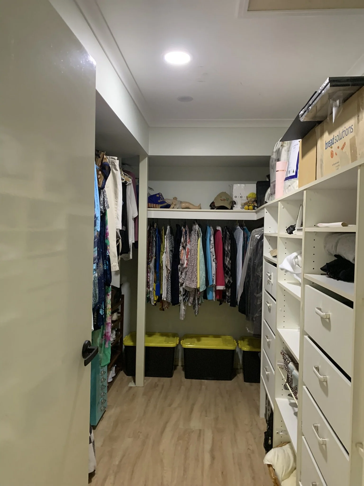 Walk-in closet with clothes hanging on racks, shelves with boxes and various items, and storage bins on the floor. The solar skylight is lighting up the walking robe.