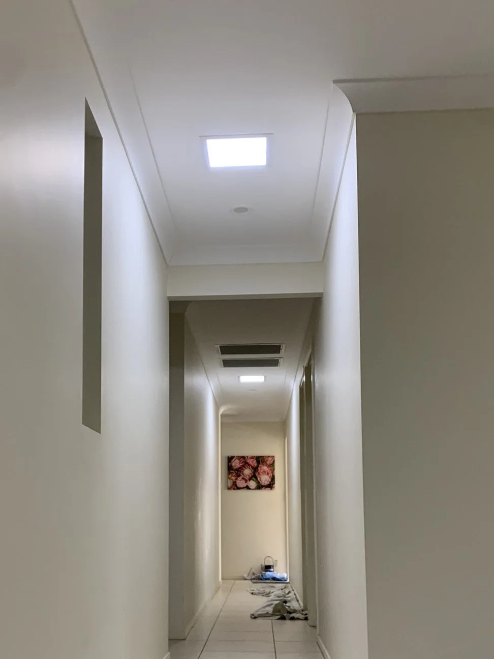 Long indoor hallway with white walls and ceiling lights, featuring a painting at the end and some items on the floor.
An illume skylight light up the space 