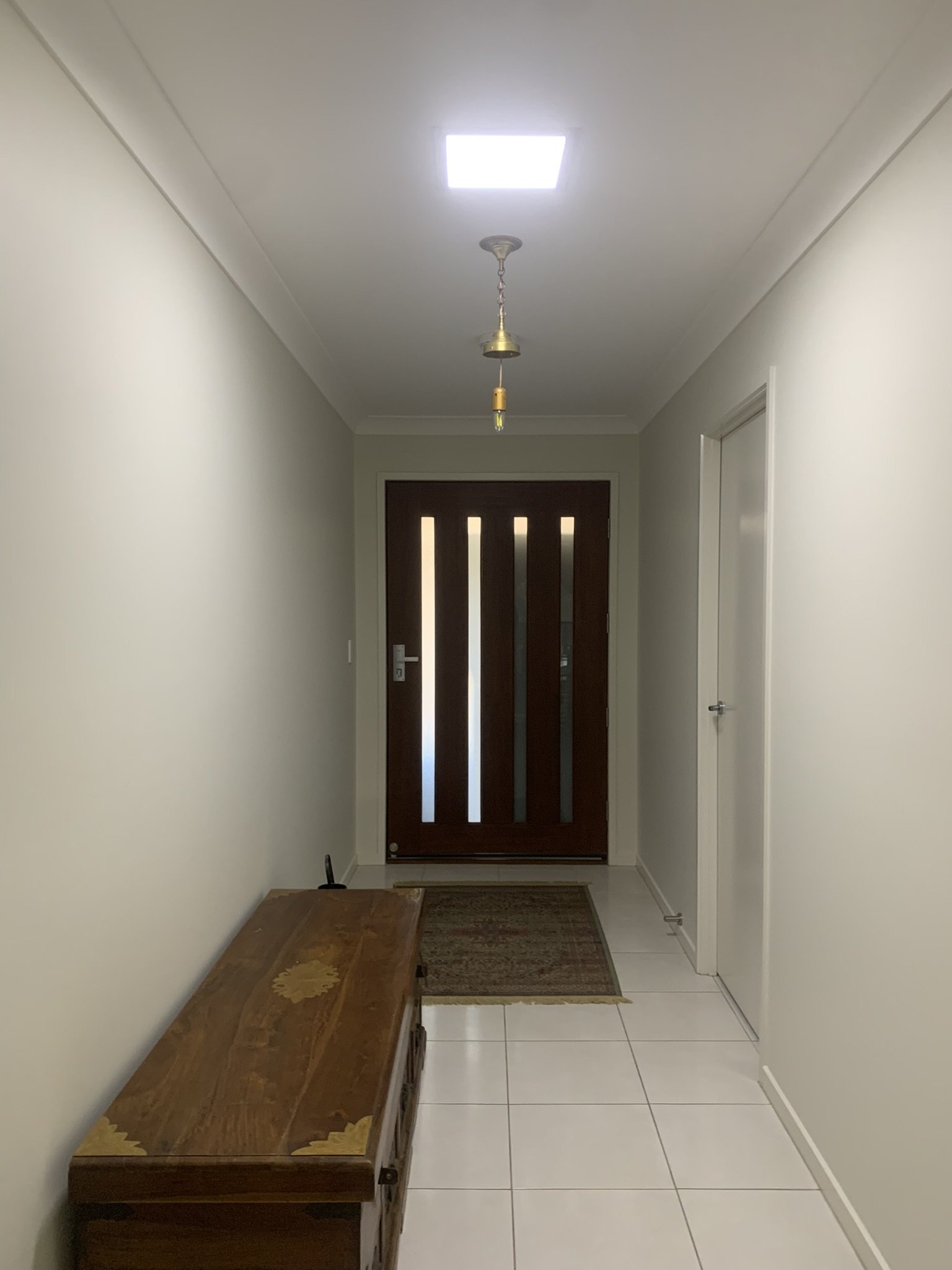 Tiled hallway with a wooden chest, leading to a modern front door, pendant light, a small floor rug and a bright illume skylight.