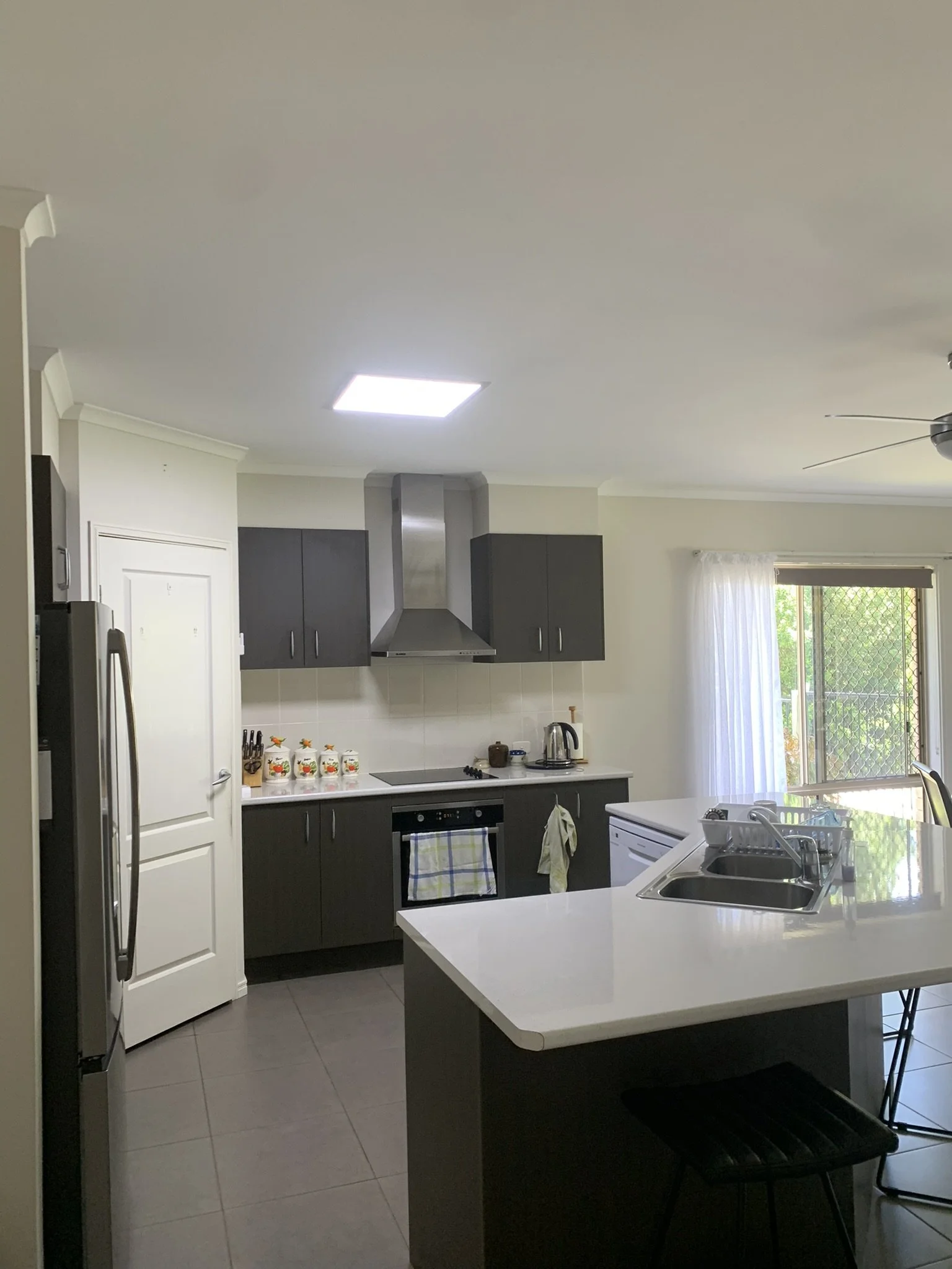 Modern kitchen with dark cabinetry, stainless steel appliances, and a light countertop. Features include an oven, stove, refrigerator, and sink on a central island. The space is bright with natural light coming through a window and an illume skylight