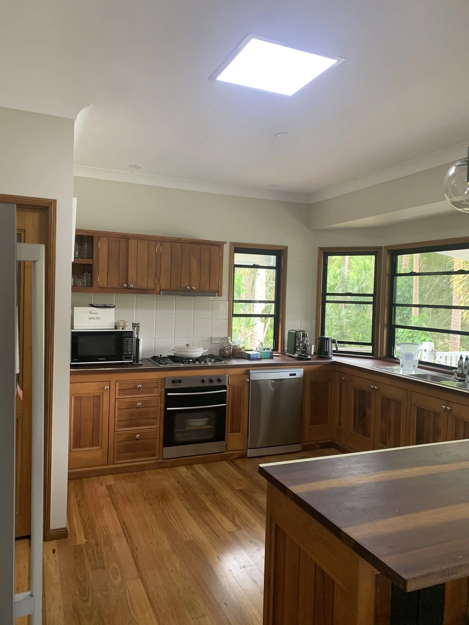 Modern kitchen with wooden cabinets and stainless steel appliances, including a stove, oven, dishwasher, and microwave. Sunlight illuminates the room through large windows. The floor is hardwood, and an illume skylight is brightening the space.