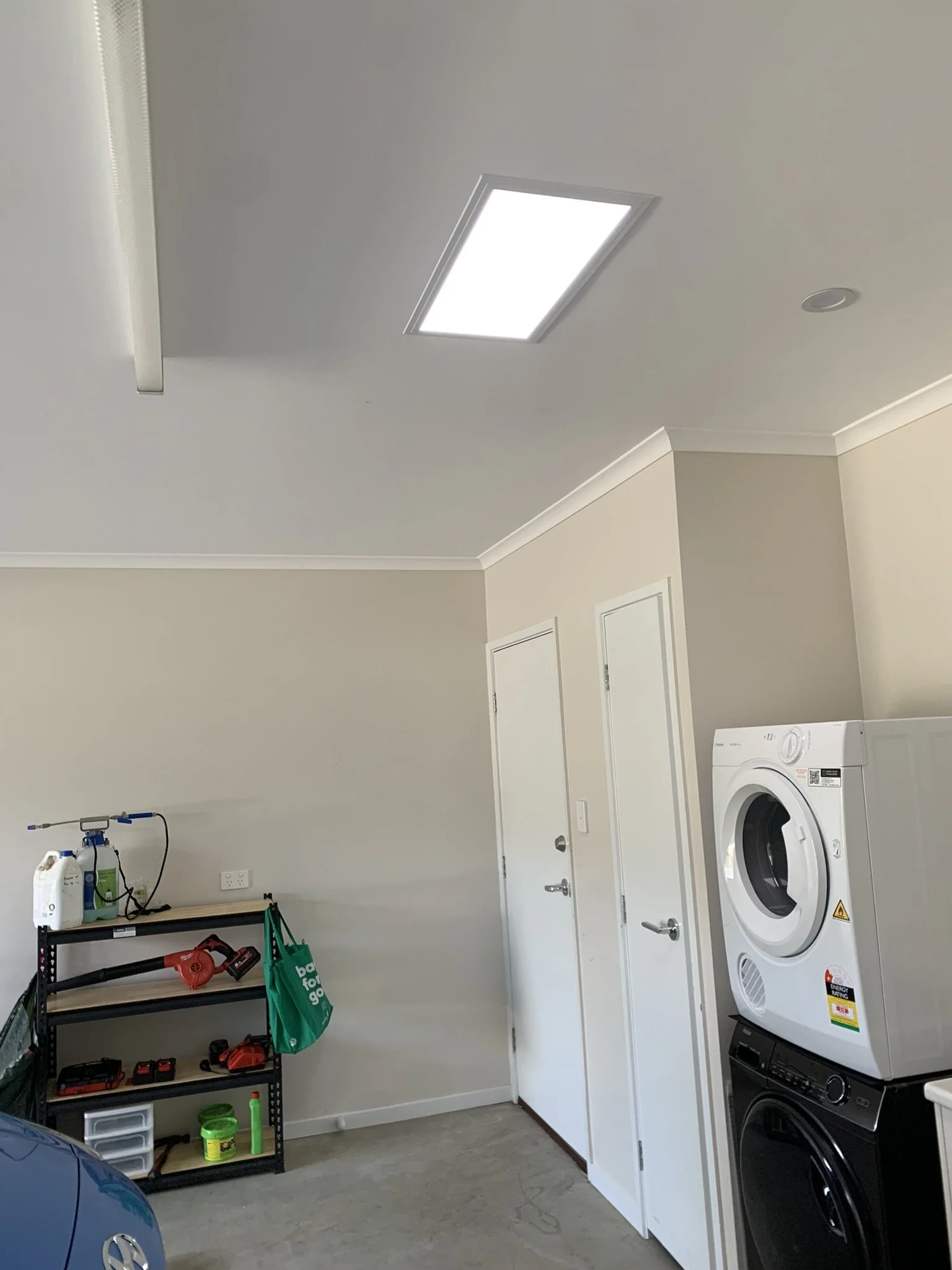 Garage interior with stacked washer and dryer, metal shelving unit, various cleaning supplies, and a blue car partially visible. A long rectangle illume skylight bring light to this space.