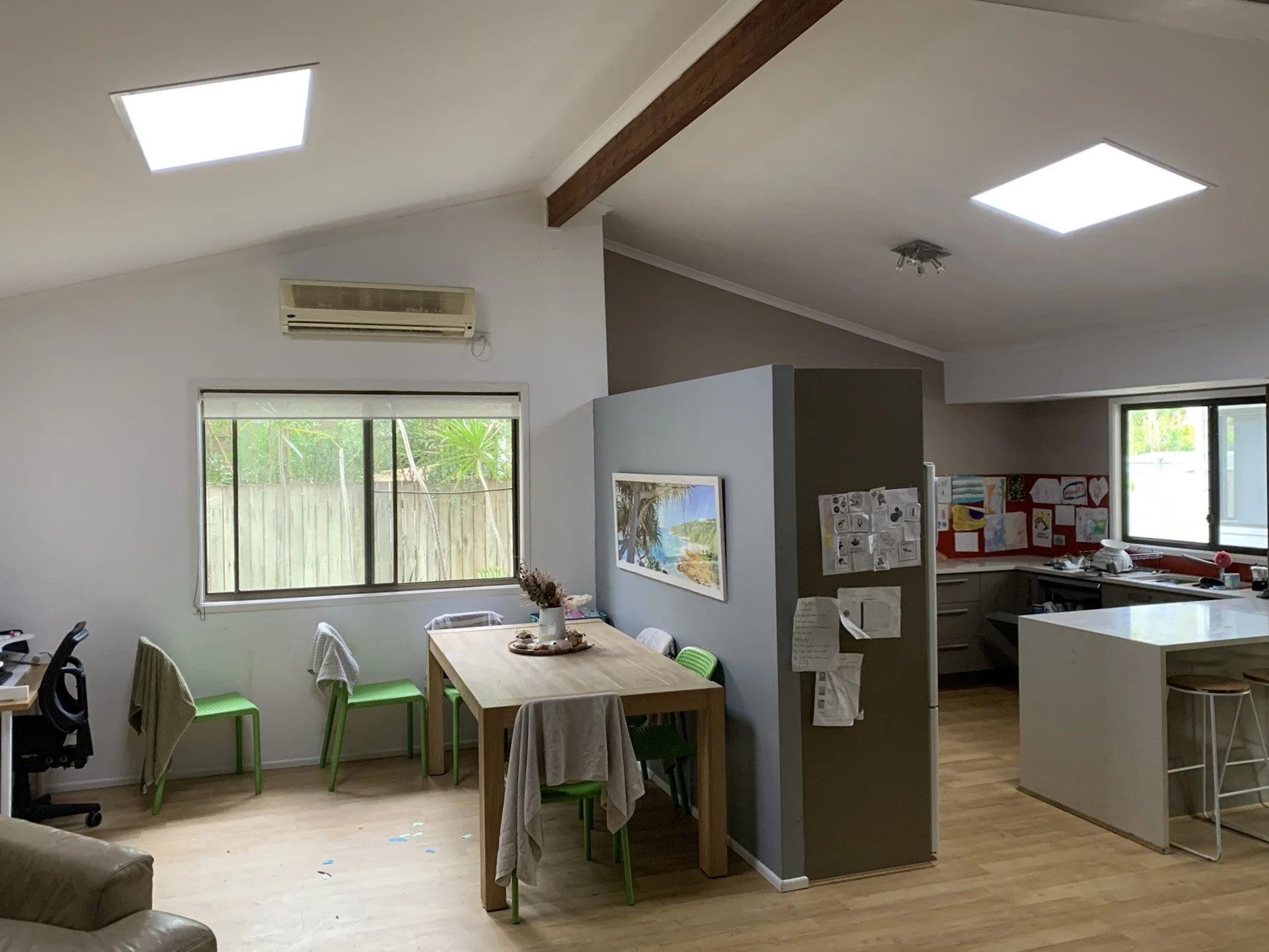 Open-plan kitchen and dining area with two illume skylights, wooden floors, a dining table with green chairs, bar stools at a kitchen island, wall art, and children's drawings on the wall.