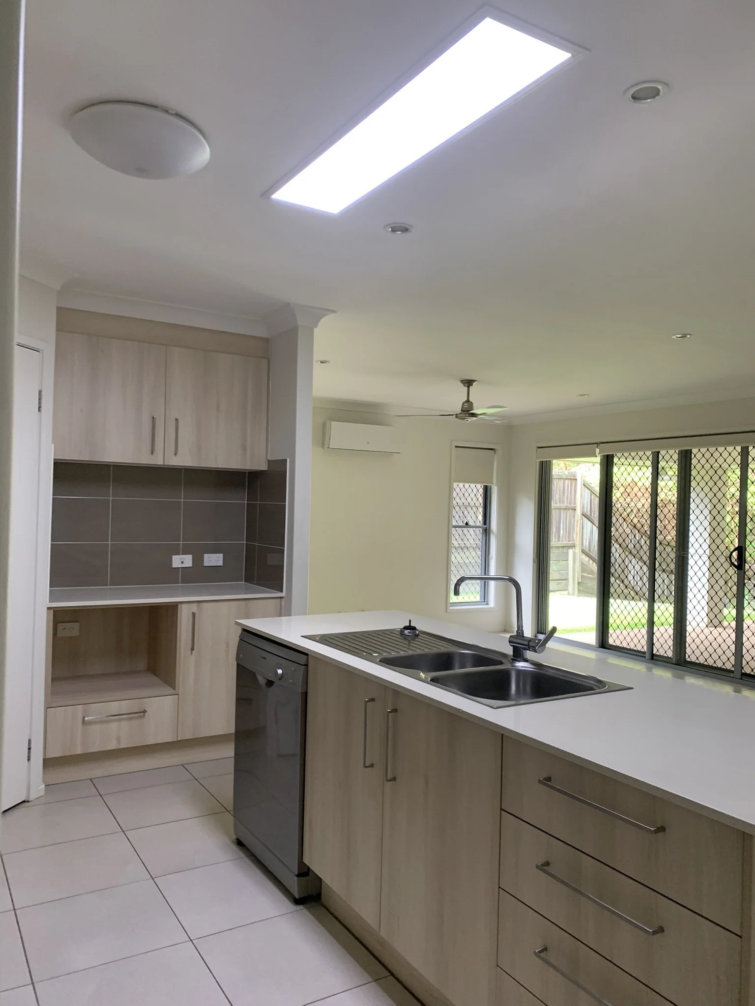 Modern kitchen with light wood cabinets, stainless steel double sink, dishwasher, gray backsplash, large window, illume skylight, and fan.
