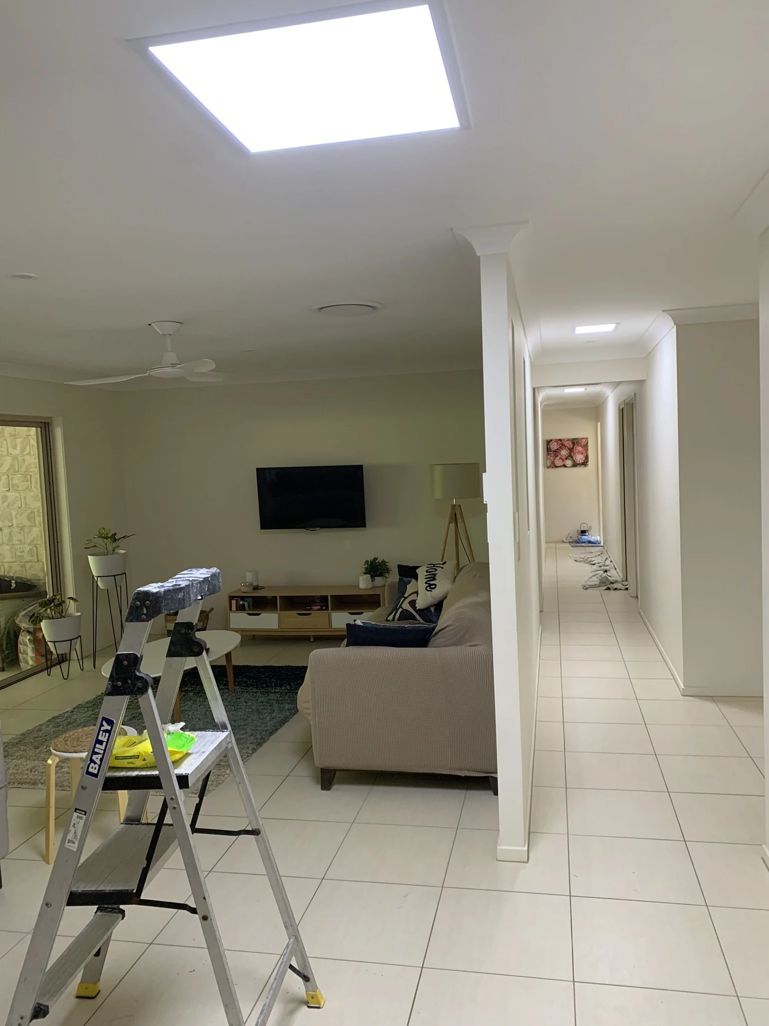 Interior of a living room with a beige sofa, wall-mounted TV, and a ceiling fan. A stepladder is in the foreground on tiled flooring. A hallway leads to other rooms, featuring patio doors some skylights