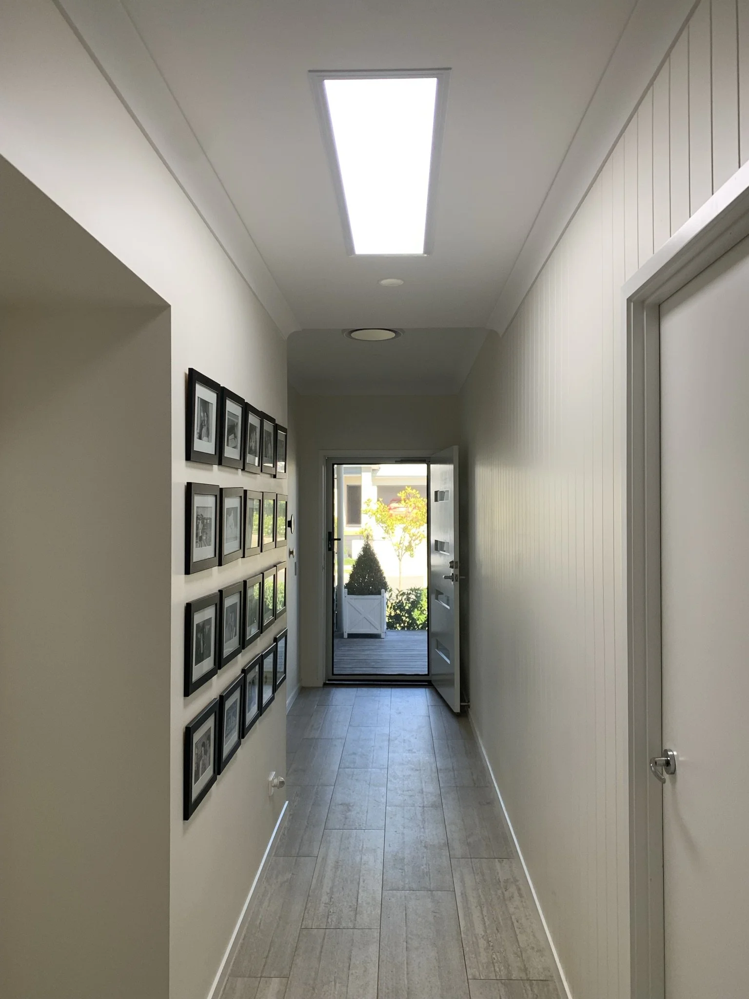 Interior hallway with wooden floor, white walls, illume skylight, photo frames on one wall, and an open door leading outside.