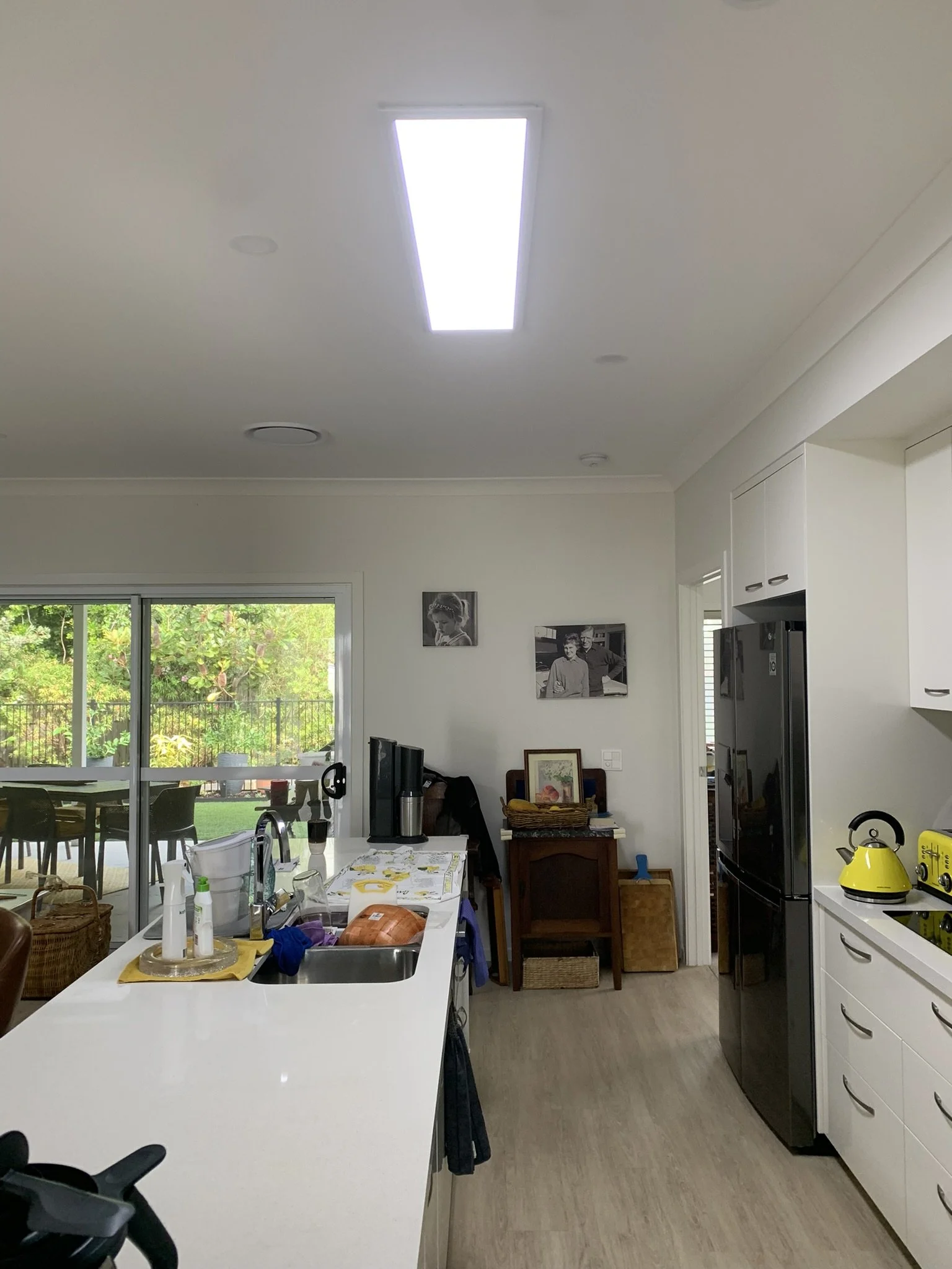 Modern kitchen with white countertops, stainless steel sink, large refrigerator, yellow kettle, and indoor plants. Light-filled space by an illume skylight with framed photos on the wall and patio view.