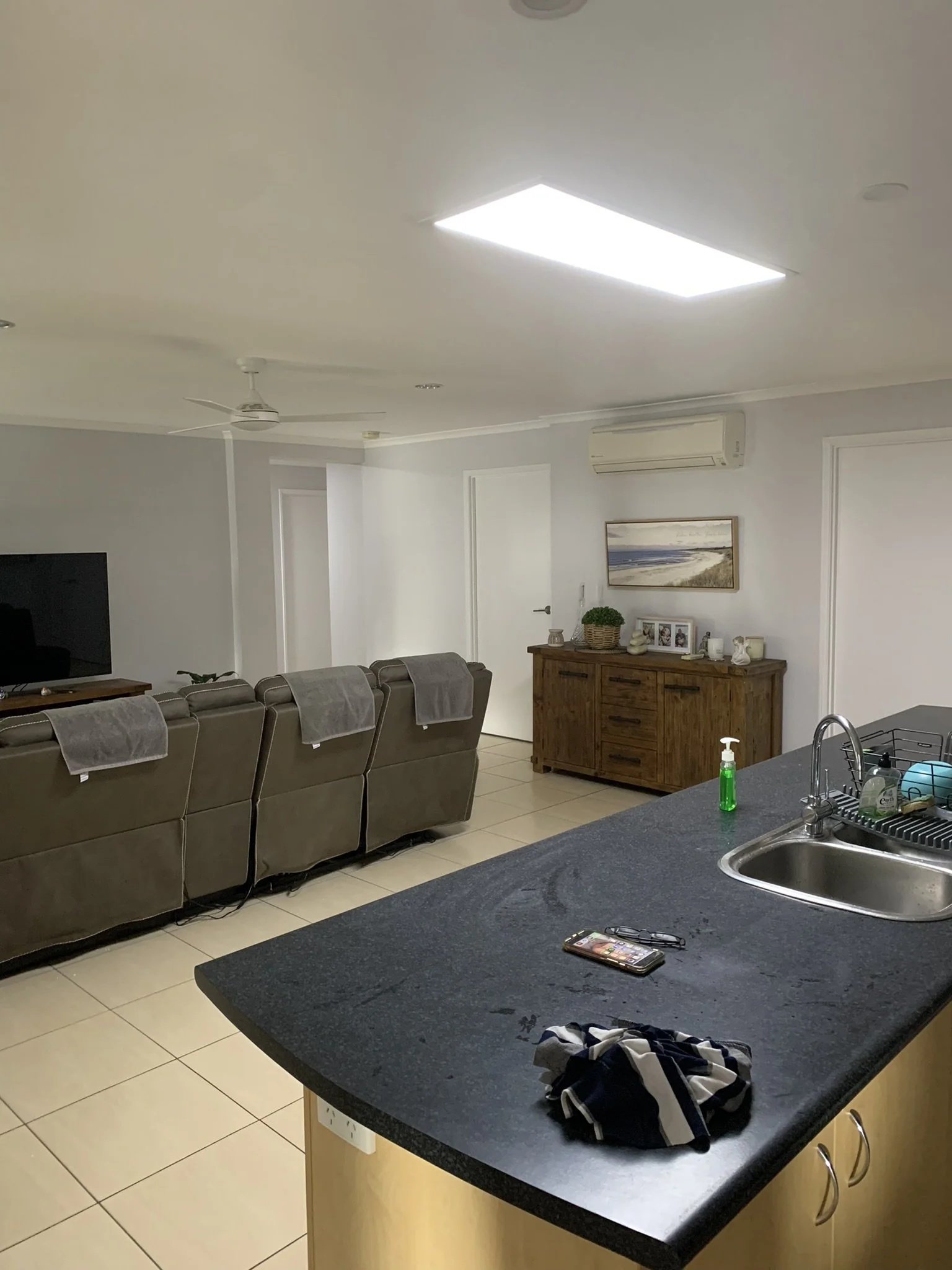 Living room with recliner sofas, an island counter with a sink, a wooden cabinet, and a TV on a stand. Towels are draped over the sofas, and various items are on the counter, including a phone and dish soap. A large skylight illuminates the space.