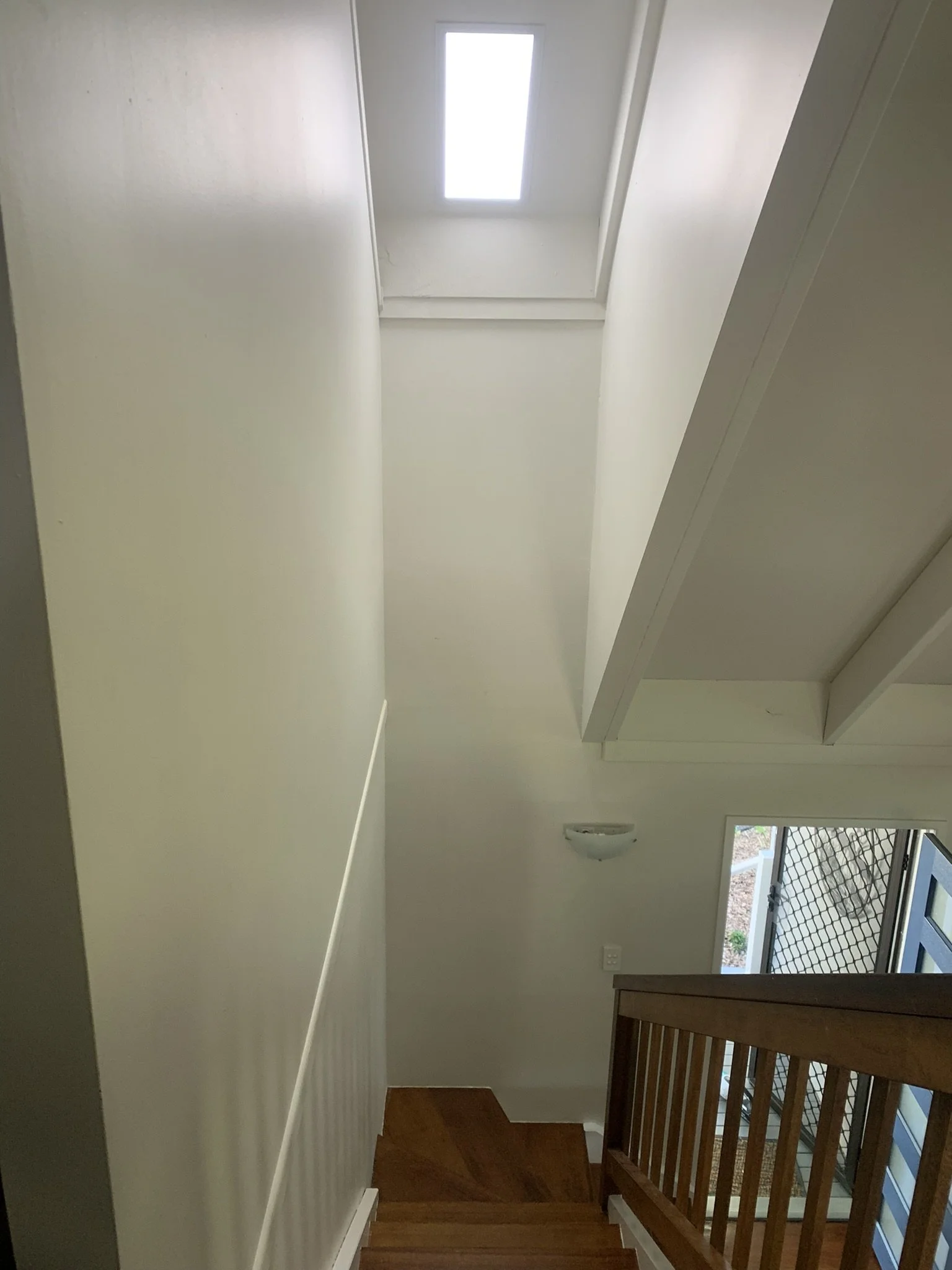 Stairwell with wooden steps and handrail, white walls, a wall-mounted light fixture, and a rectangular skylight on the ceiling.
