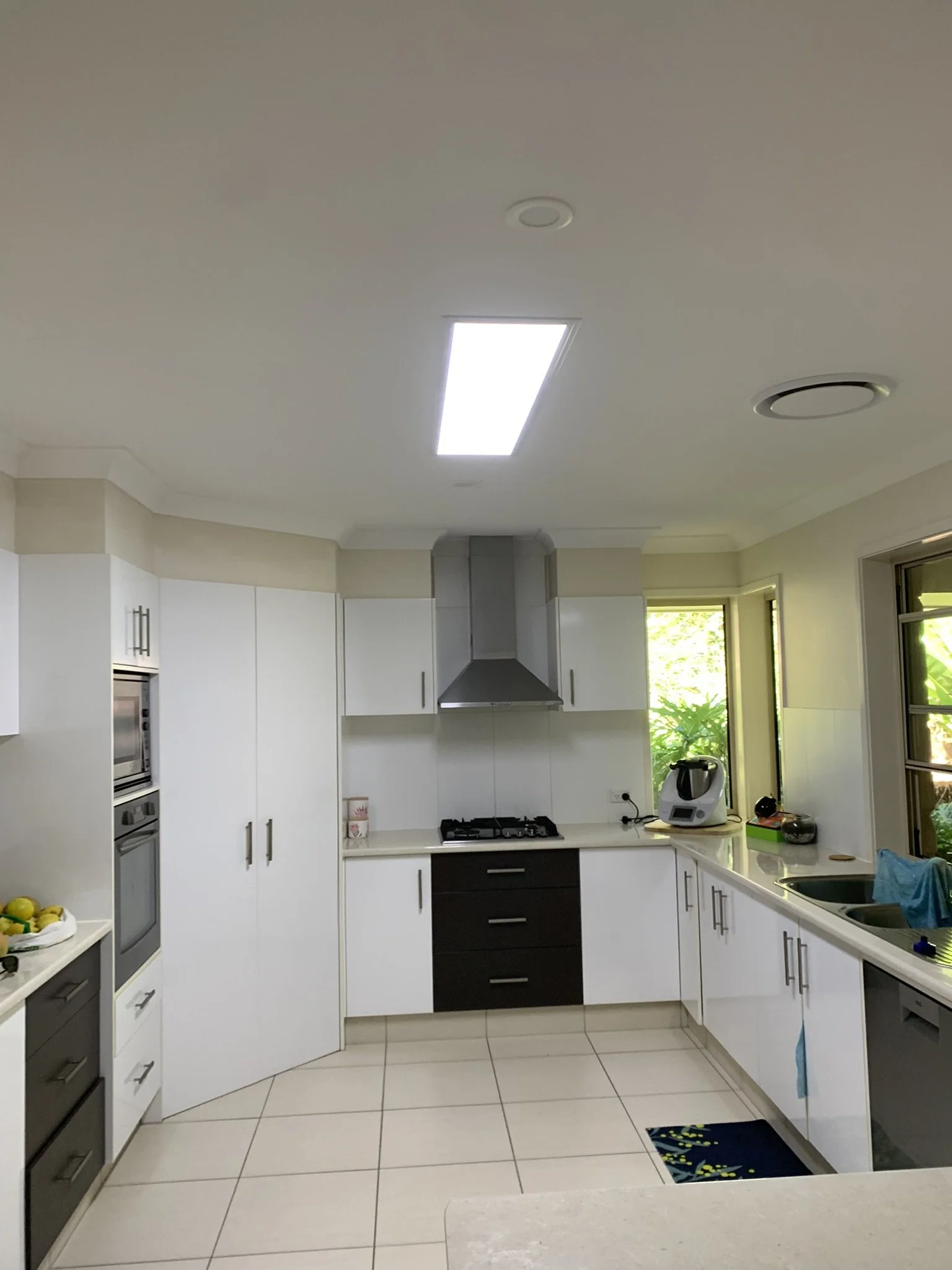 Modern kitchen with white cabinets, tile flooring, stainless steel appliances, and a large window. The kitchen features a range hood, oven, and a countertop with a bowl of fruit and a small appliance. The illume skylight bring light in the space.