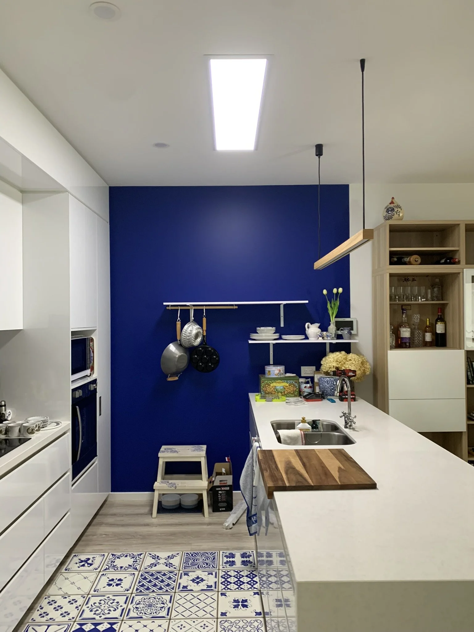 Modern kitchen with blue accent wall, white cabinetry, illume skylight, and patterned floor tiles. Features a countertop with sink, wooden cutting board, and various kitchen items.