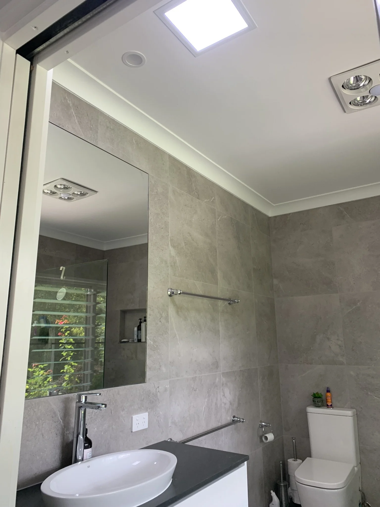 Modern bathroom with gray tile walls, a white sink on a dark countertop, a large mirror, towel racks, a toilet, and natural light from an illume skylight.