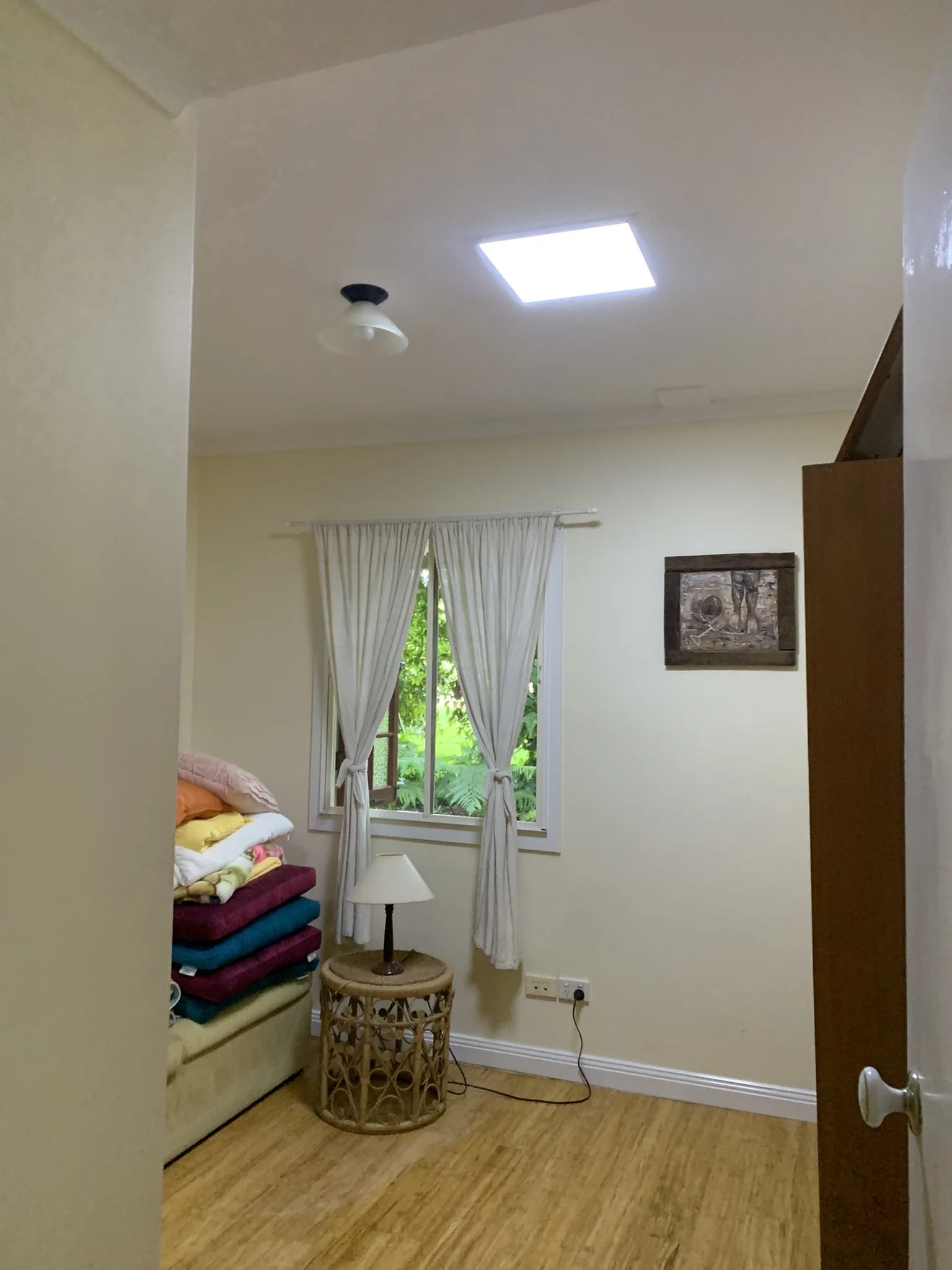Small room with light-colored walls, a window with white curtains, and a round wicker table with a lamp. A stack of folded blankets or towels is on a beige sofa to the left. Ceiling features a light fixture and skylight, and there's a decorative wall