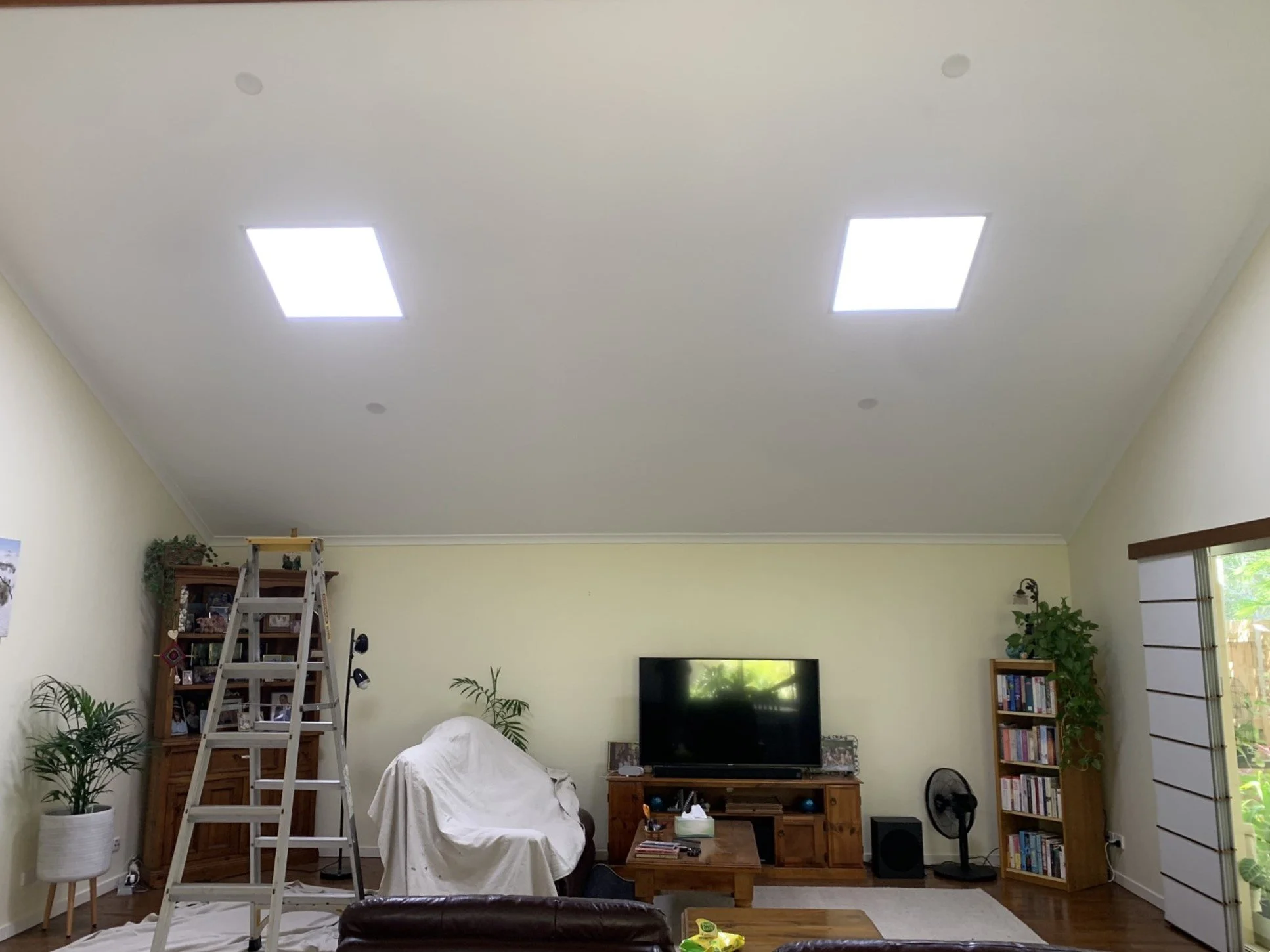 Living room with ladder, covered furniture, bookshelves, TV, and illume skylights on the ceiling.