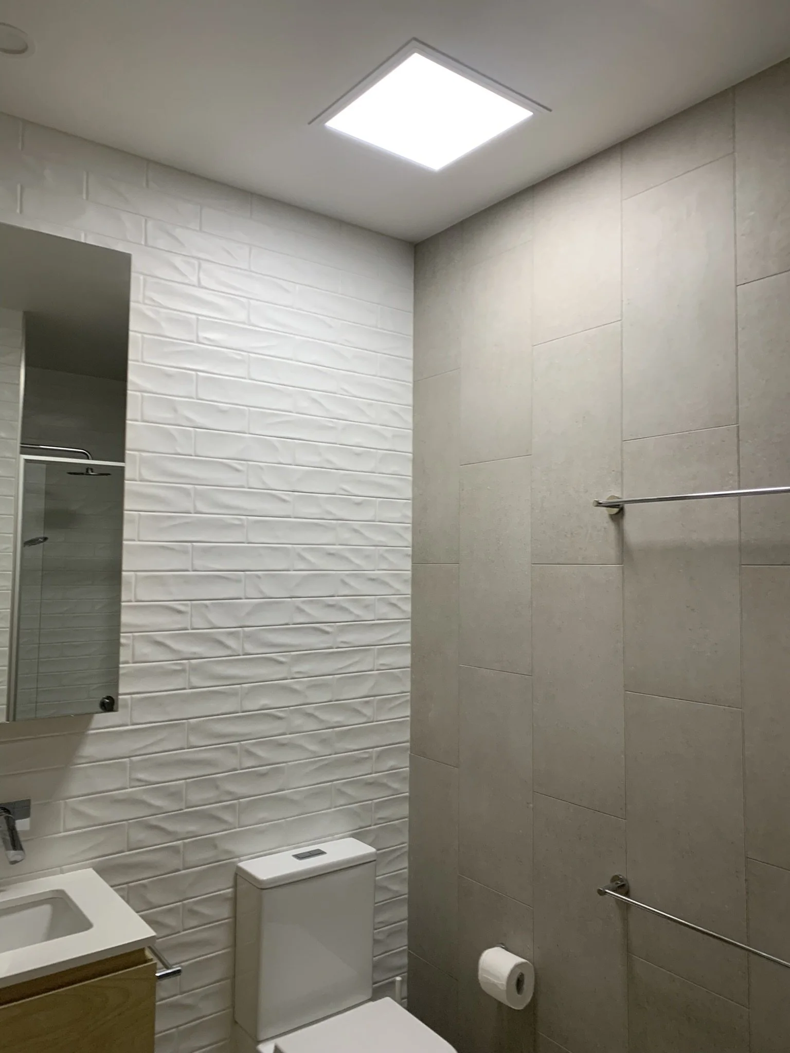 Modern bathroom with a white toilet, wall-mounted sink, large mirror, white textured tile on one wall, gray tile on the other, and a skylight.