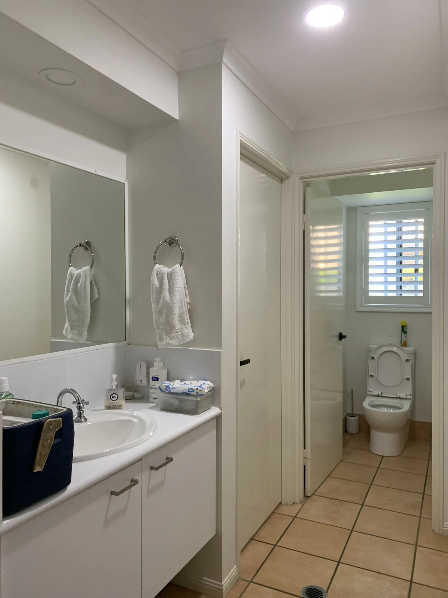 Bathroom with sink, mirror, and toilet; white towels hanging on rack, toiletries on counter, tiled floor. An illume skylight  is making the space look warm