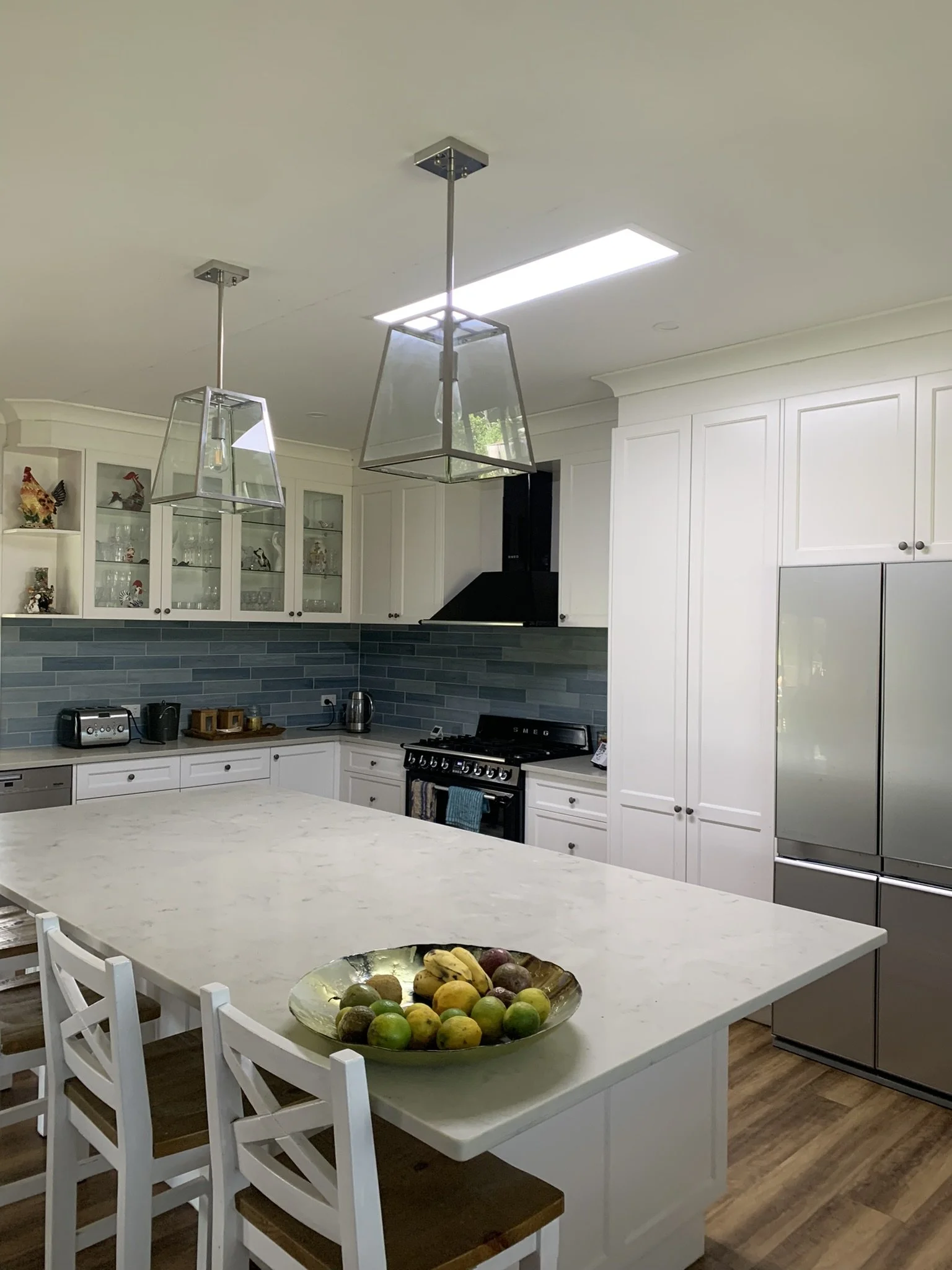 Modern kitchen with white cabinets, a gray tile backsplash, and stainless steel appliances. The room features a large kitchen island with white stools. Overhead lighting includes an illume skylight and pendant lights. Wooden flooring.