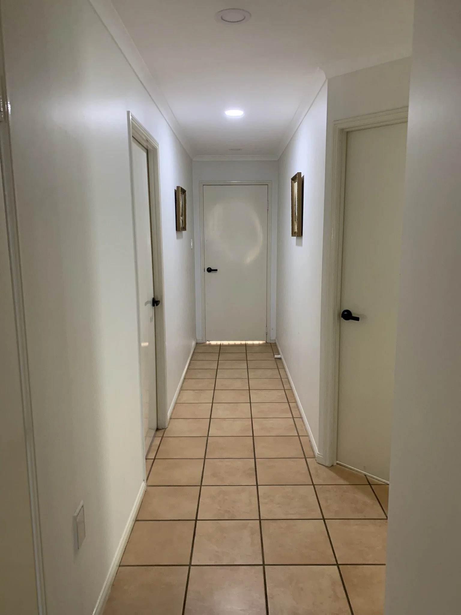 Long hallway with beige tiled floor, white walls, and several closed doors. An illume skylight is lighting up the corridor. 