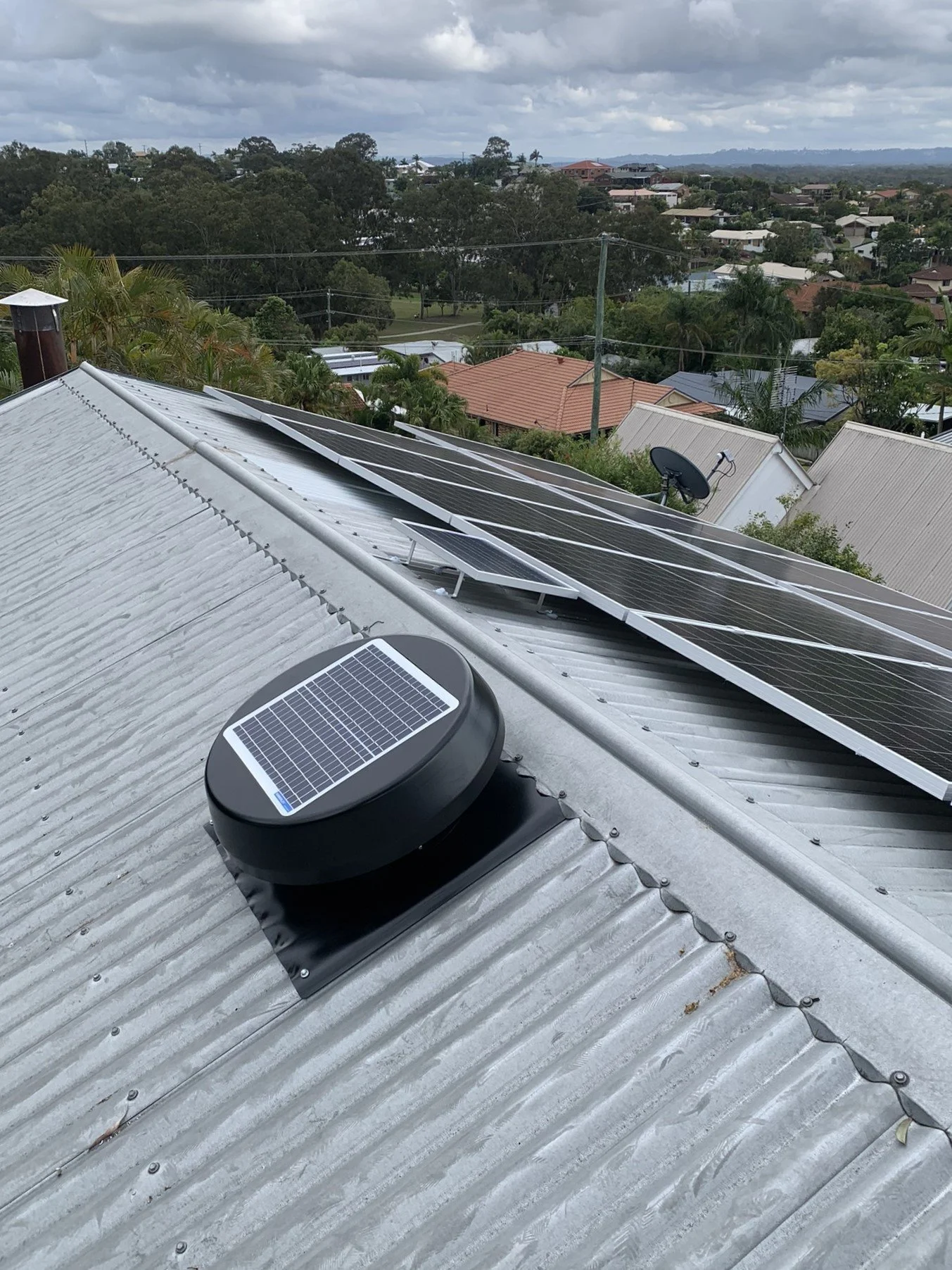 Metal roof with solar panels and a solar-powered ventilation fan (KSV300), overlooking a suburban area with trees and houses.