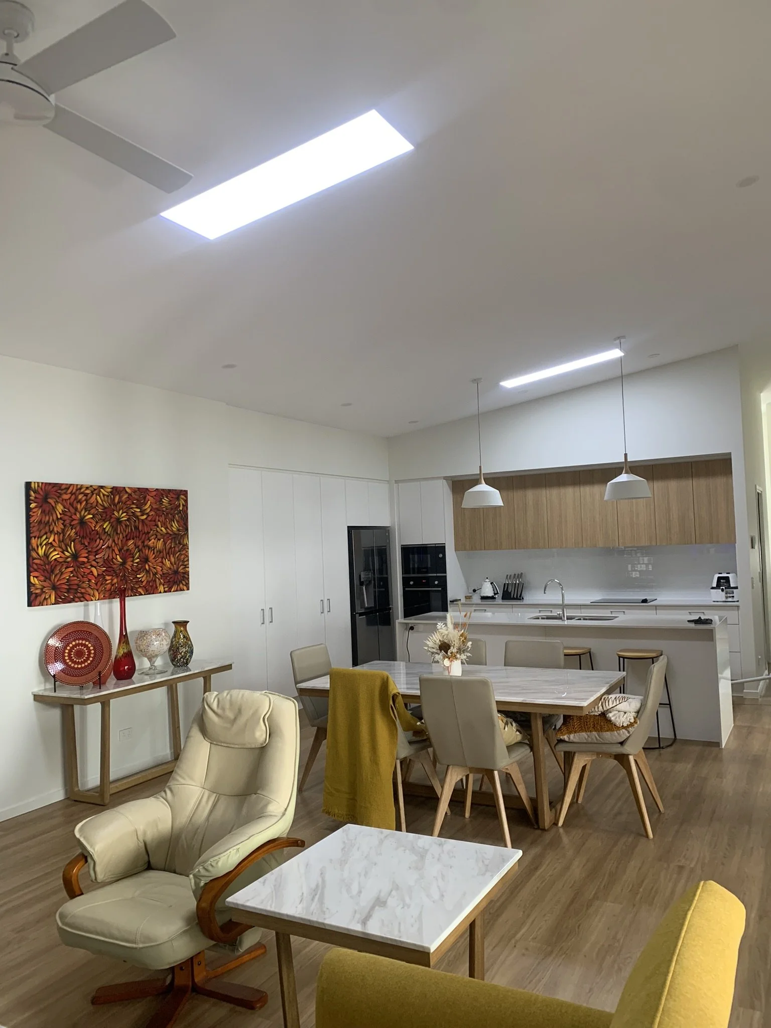Modern open-plan kitchen and dining area with illume skylights, white cabinets, wood accents, and a marble dining table with beige chairs. A comfortable seating area features a cream armchair and small table. 