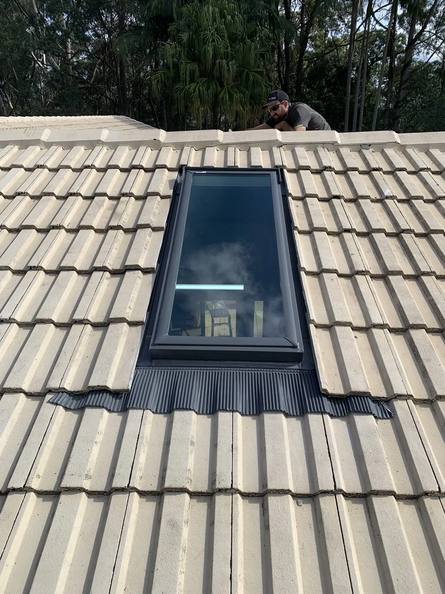 Roof with a skylight window surrounded by beige tiles, with a person working near the roof's edge, trees in the background.