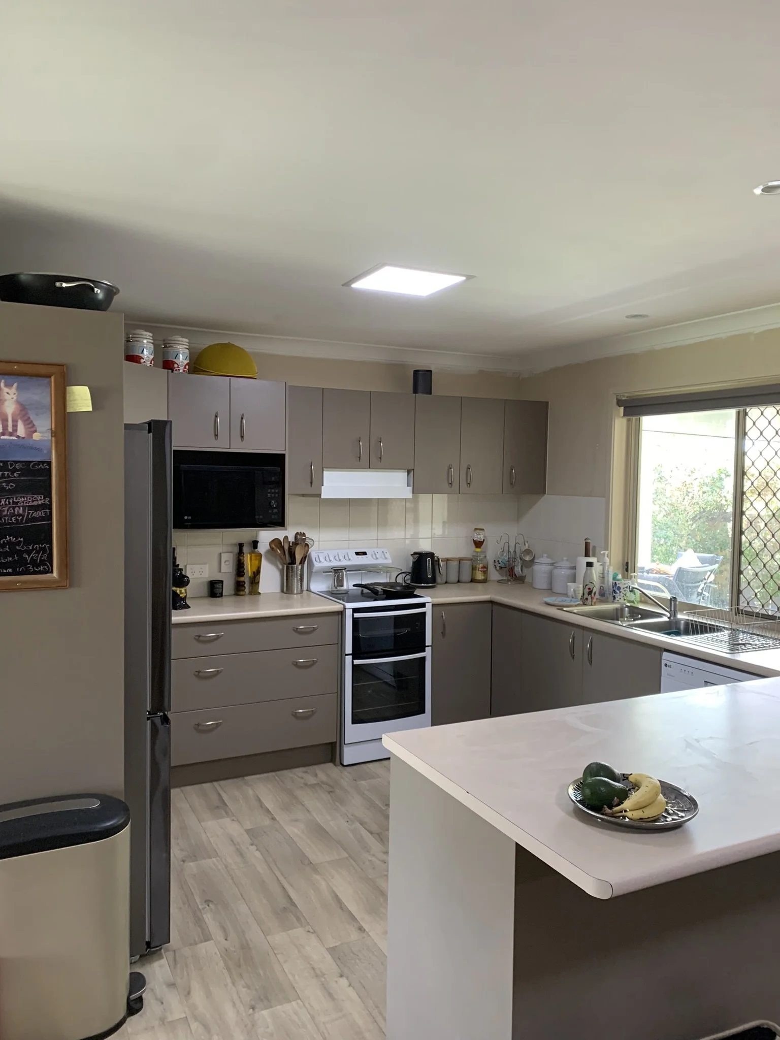 Modern kitchen with beige cabinets, stainless steel appliances, and a countertop with fruit. There is a window, a sink, various kitchen utensils on the counter, and a door leading outside and a bright solar skylight from illume.