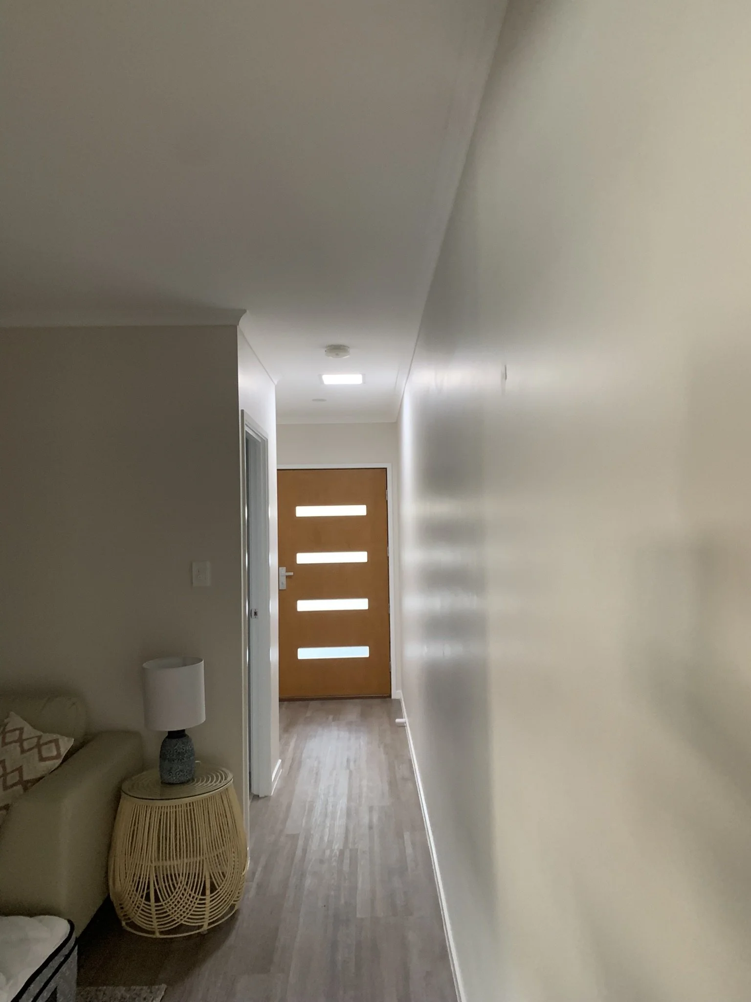 A hallway leading to a wooden front door with horizontal glass panels. There is a round woven side table with a lamp next to a beige sofa on the left. The floor is made of wood, and the walls are painted white. A small illume skylight bring light.