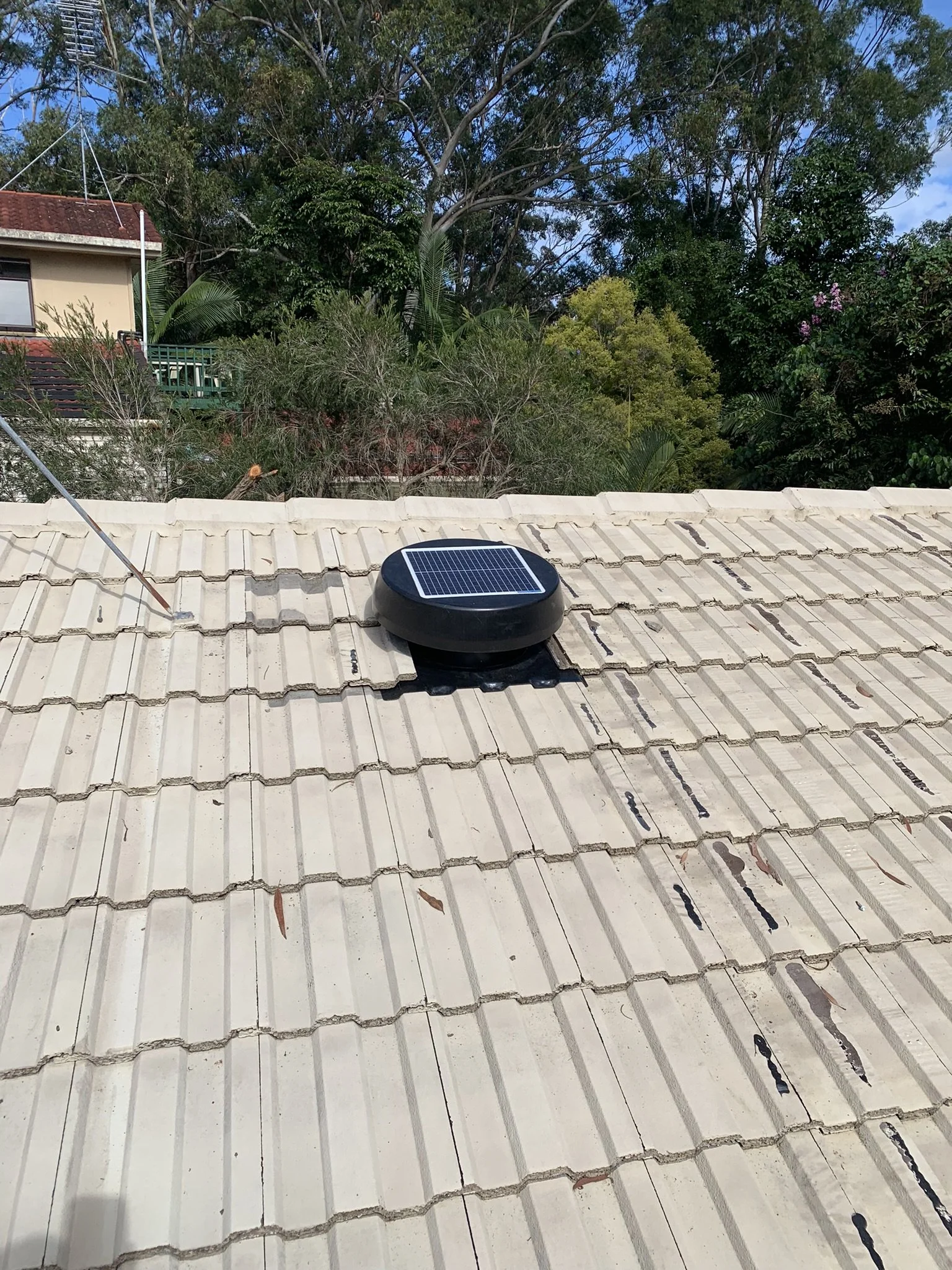 A solar attic fan installed on a tiled roof with surrounding trees and neighboring houses in the background.