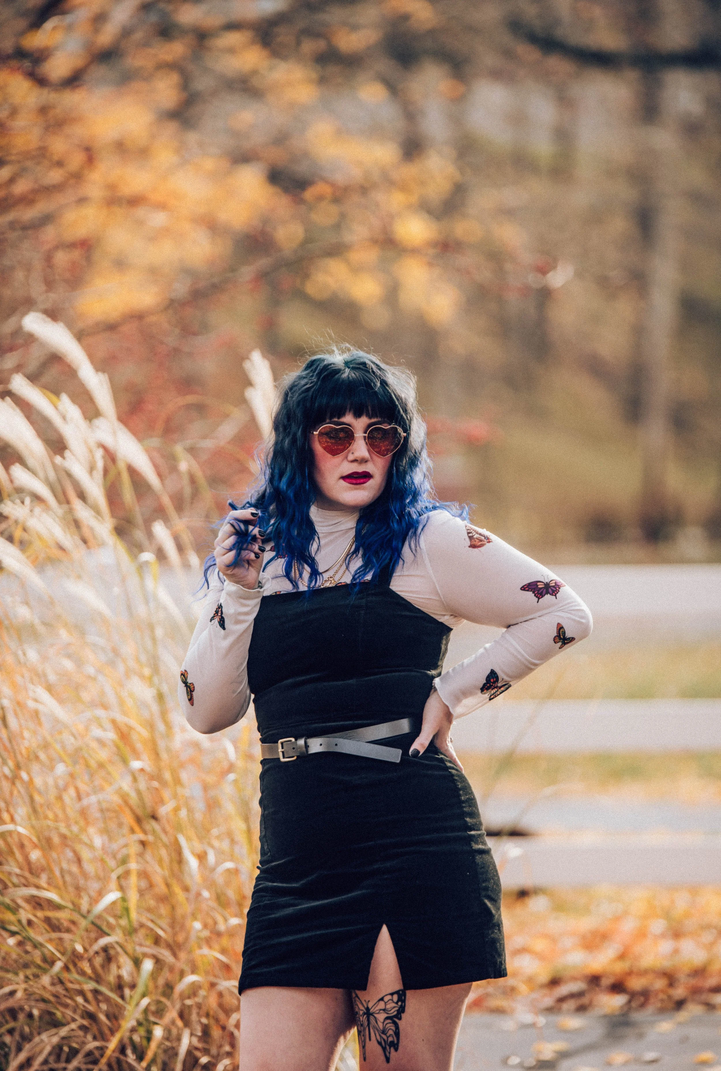 A woman with blue and black wavy hair standing outdoors among fall foliage, wearing sunglasses, a butterfly-printed long sleeve shirt, a black overall dress with a slit, and a white belt.