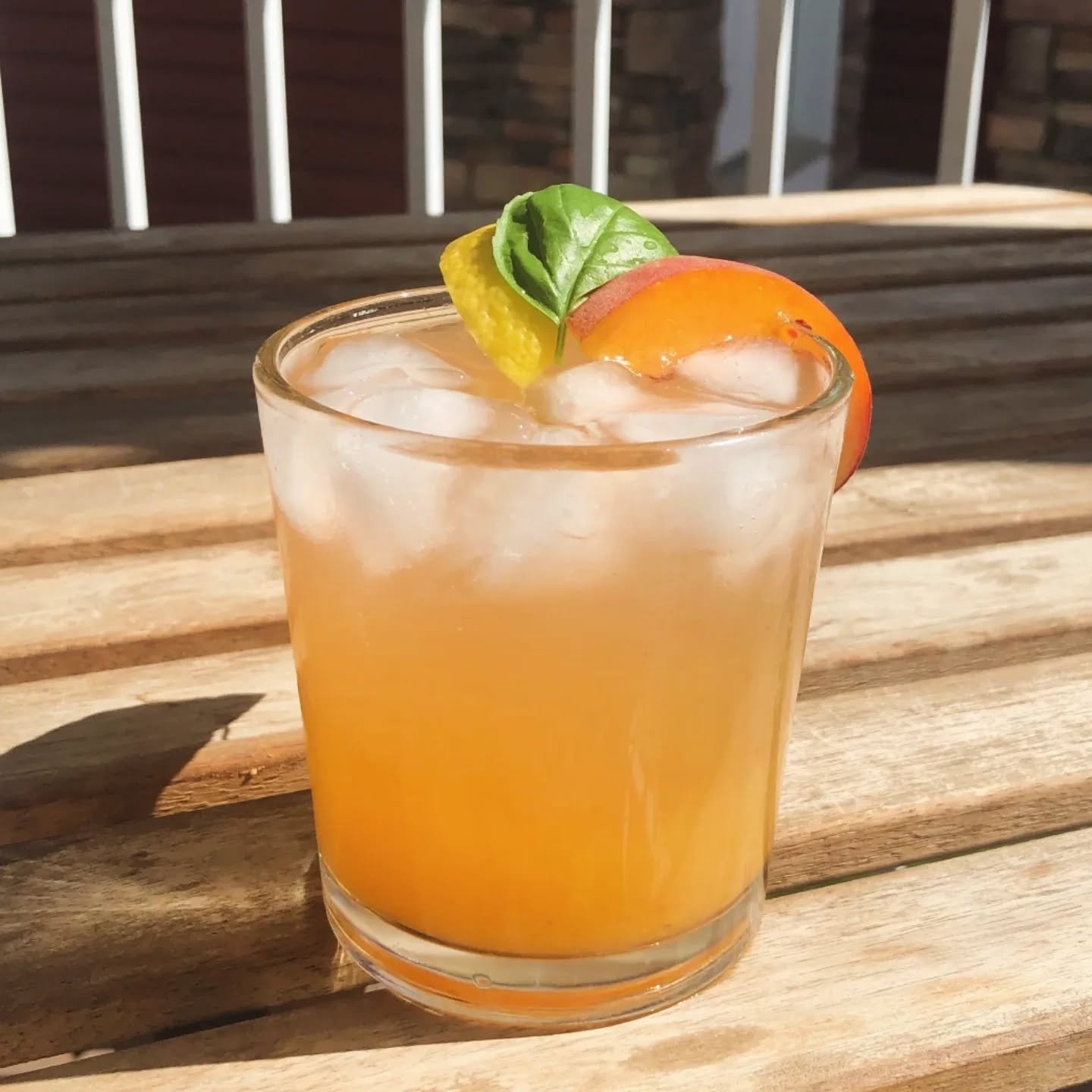 A refreshing cocktail in a glass with ice, garnished with a lemon slice, orange slice, and a basil leaf, placed on a wooden table.