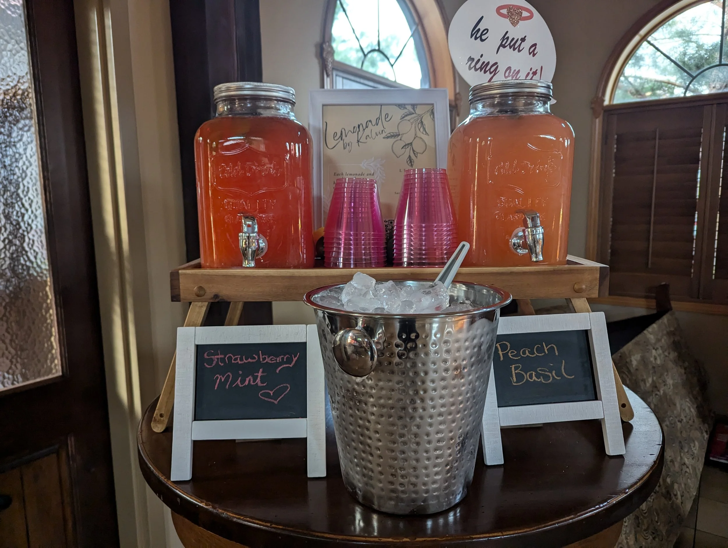 A refreshment station with dispensers labeled "Strawberry Mint" and "Peach Basil," accompanied by a bucket of ice and pink cups.