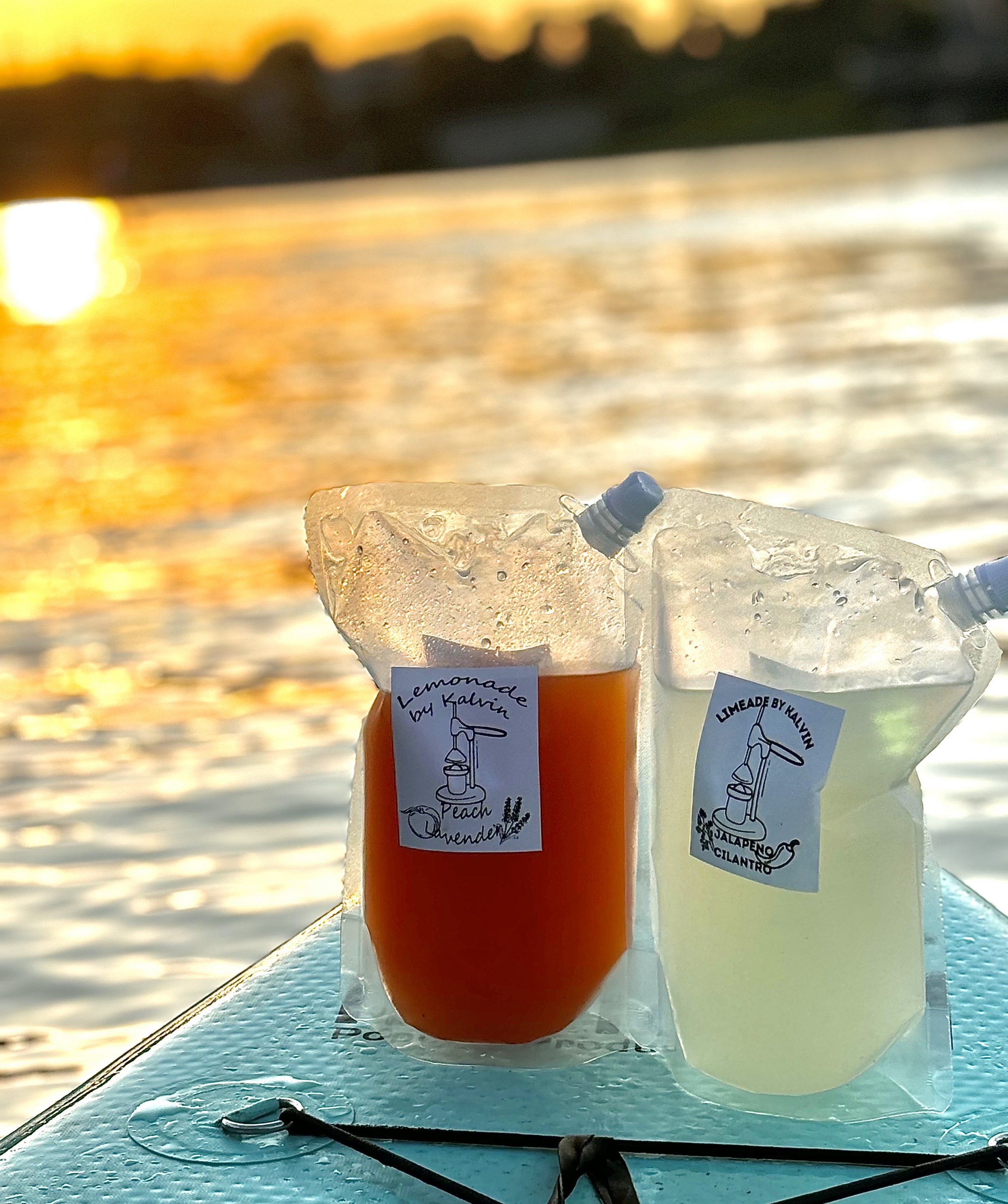 Two pouches on a paddleboard holding peach lavender lemonade and jalapeño cilantro limeade with a sunlit water view.