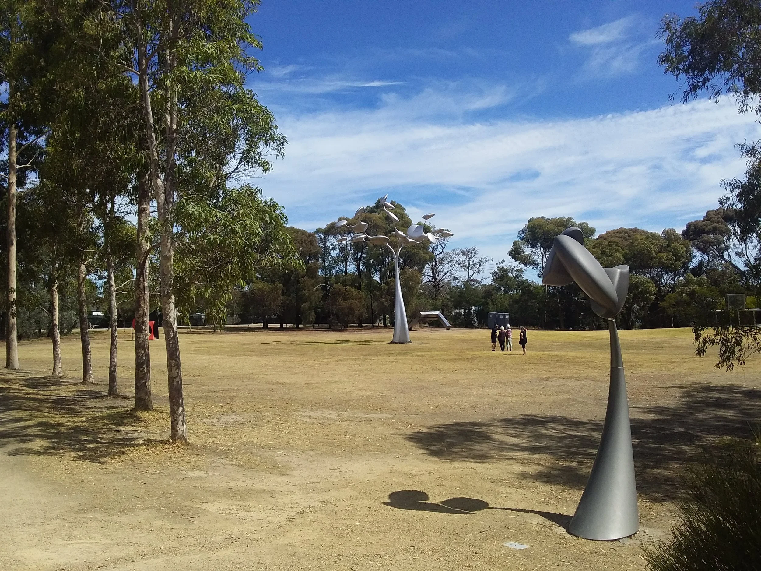 Park landscape with abstract metal sculptures, several trees, and people walking in the background under a blue sky.