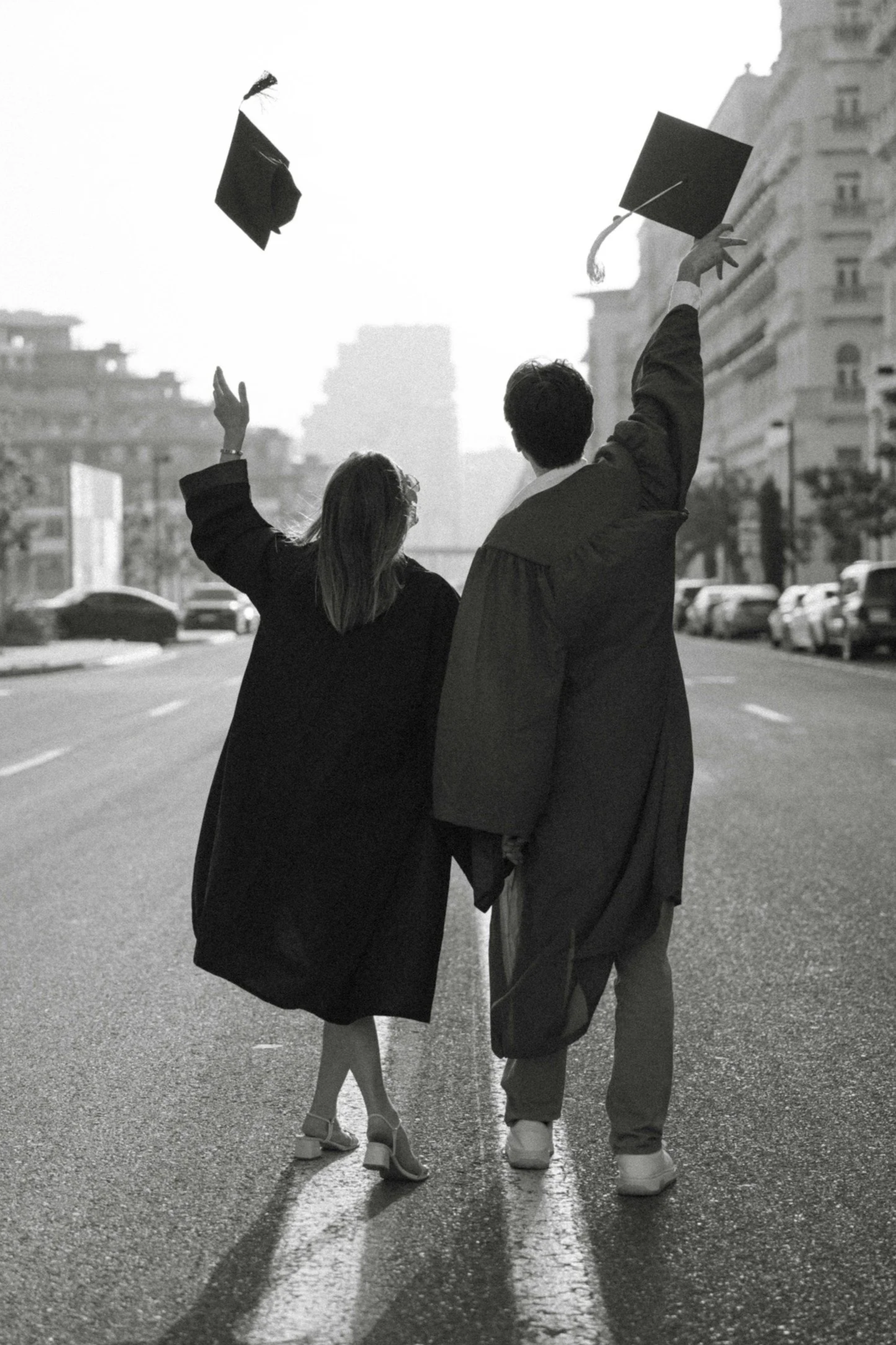 Two people in graduation gowns and caps are walking on a city street, celebrating their graduation by tossing their caps in the air.