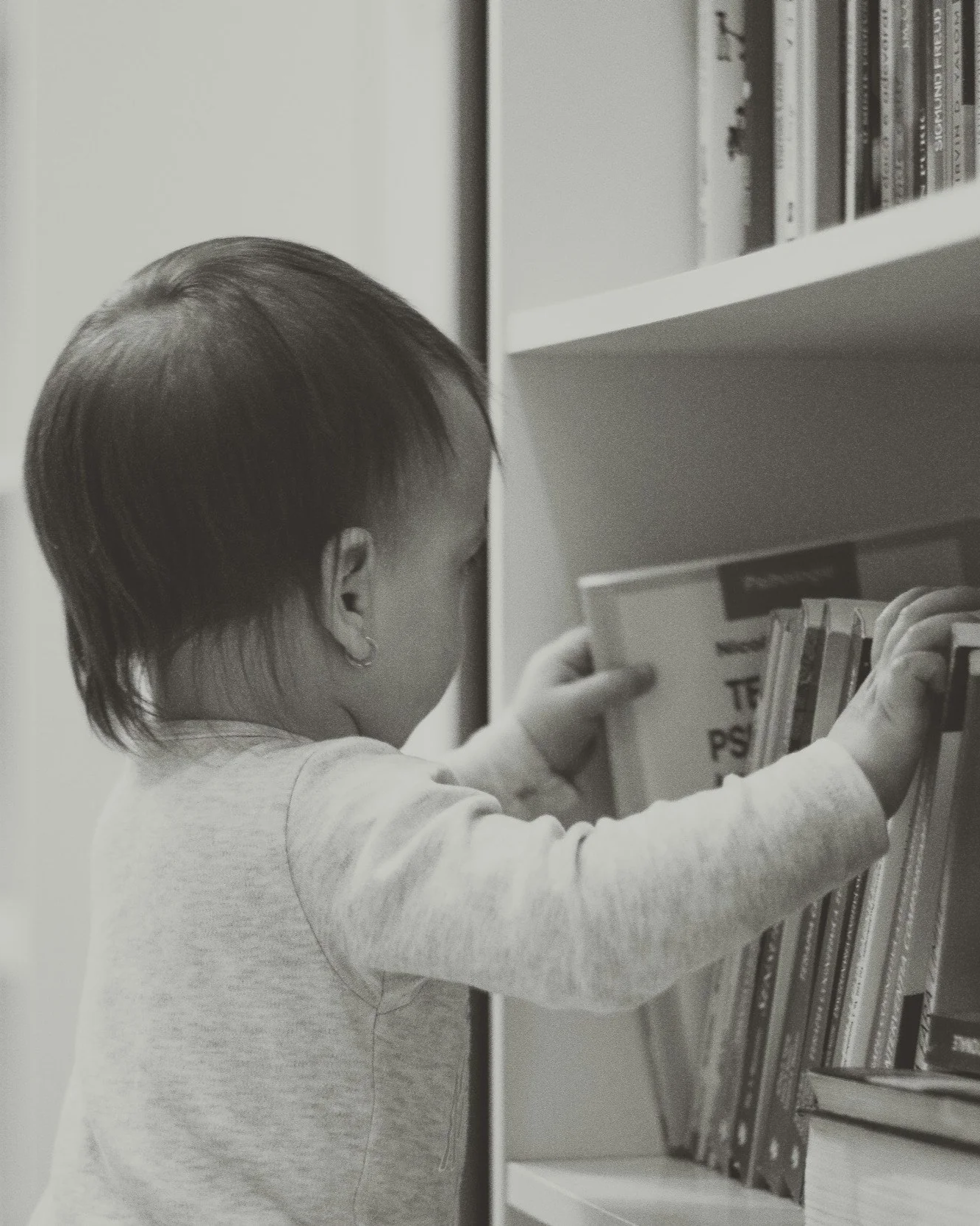 A young child with short dark hair and wearing a light-colored long sleeve shirt shelves books in a white bookcase.