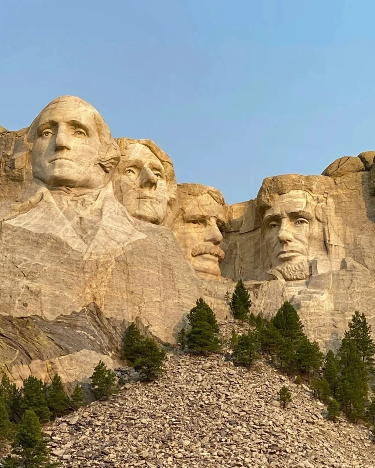 Close-up of Mount Rushmore featuring the carved faces of four American presidents against a clear blue sky, with pine trees at the base.