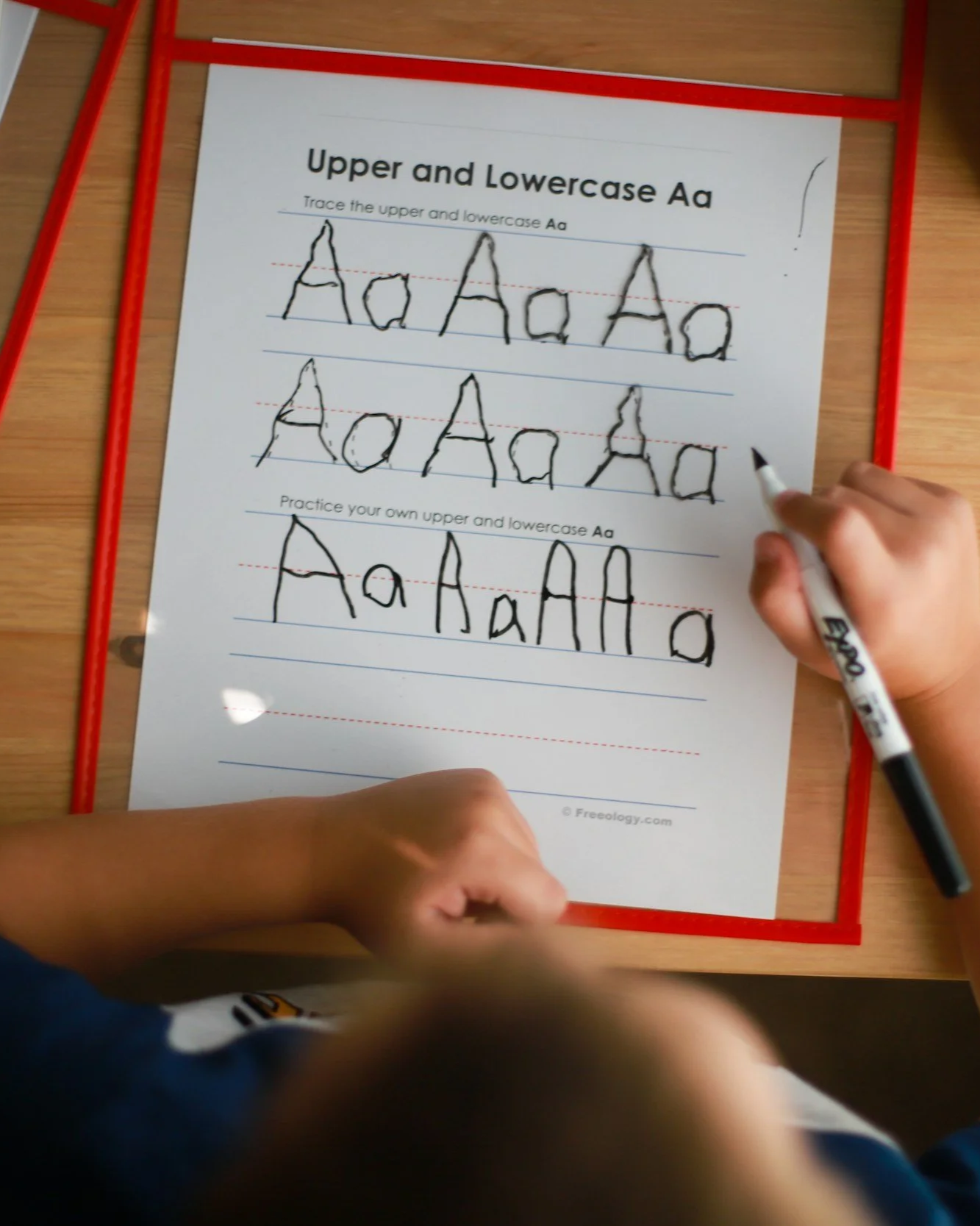 A child's hands writing on a worksheet that teaches uppercase and lowercase letter A in black marker, with a red border, on a wooden desk.