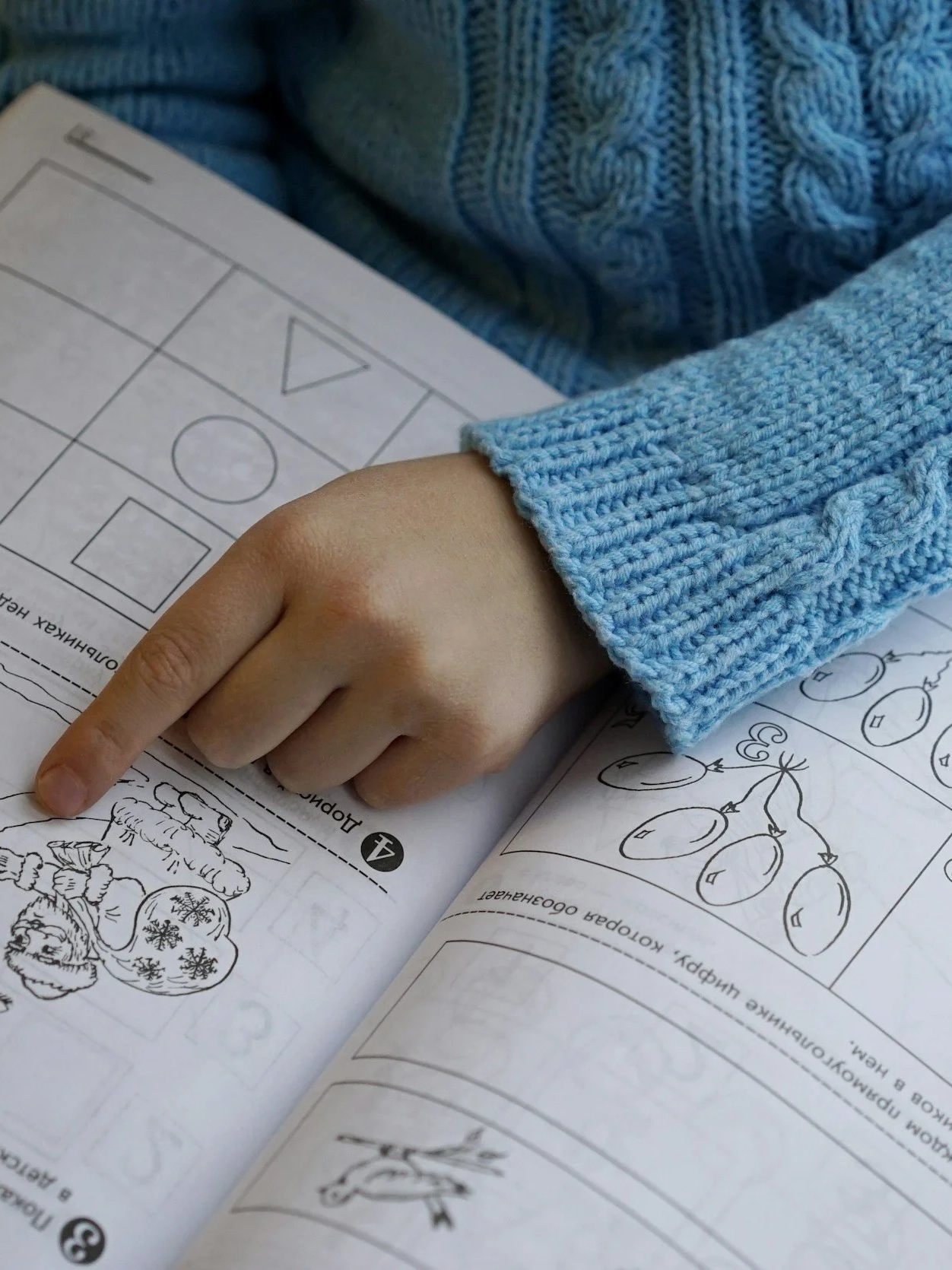 A child wearing a blue knitted sweater points to a coloring page with Christmas-themed images and holiday activities.