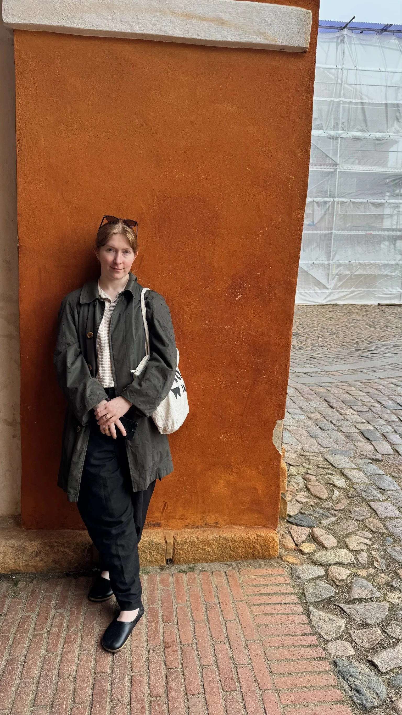 A young woman stands against an orange wall with a textured surface. She is wearing a dark raincoat, black pants, and black shoes, holding a phone in her right hand. She has sunglasses resting on her head and carries a white bag over her left shoulder. The ground consists of brick and cobblestone paving, with a construction site covered in white plastic sheeting visible in the background.